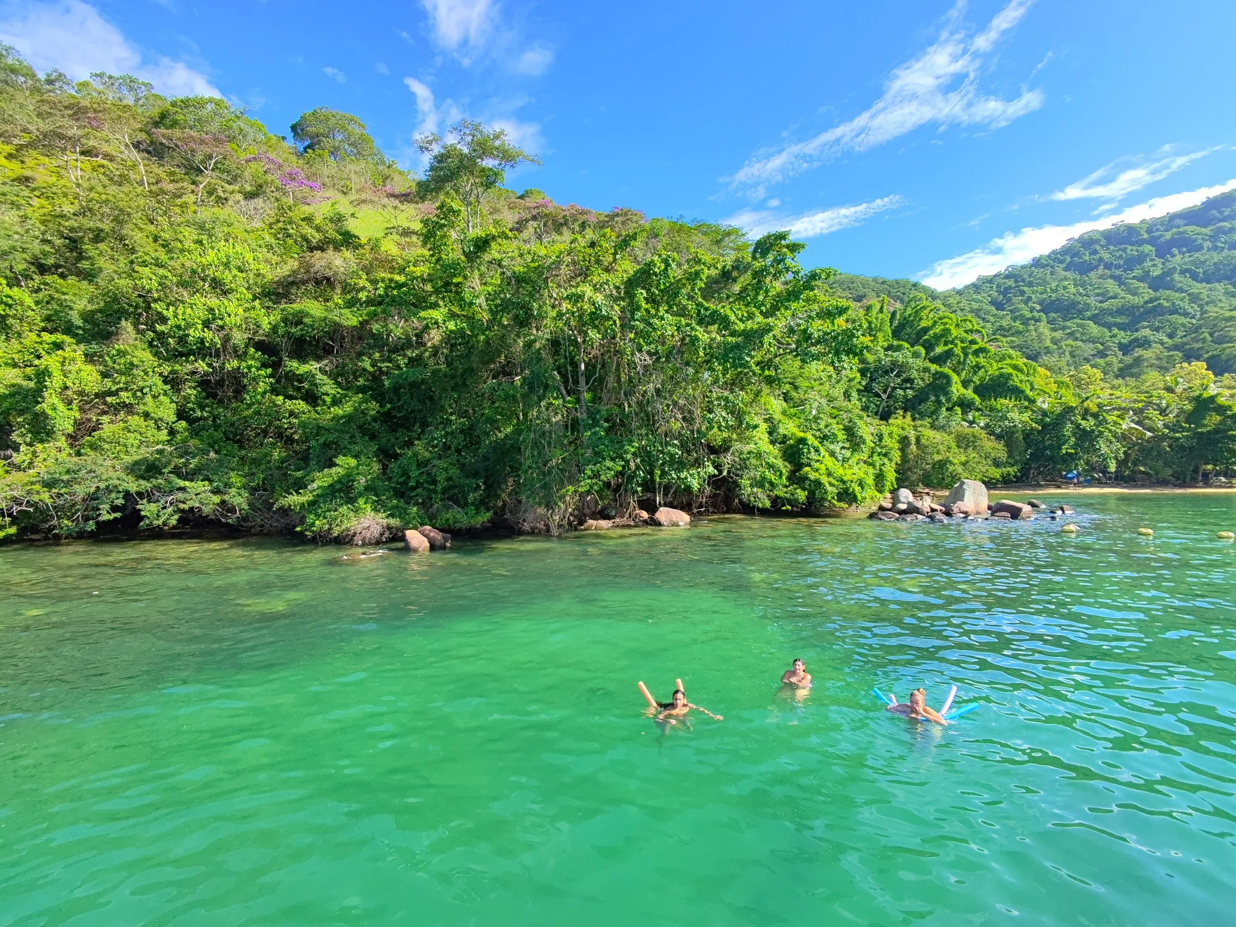 People swimming in a clear green body of water near a lush, green hillside with trees and shrubs, and a blue sky with scattered clouds.