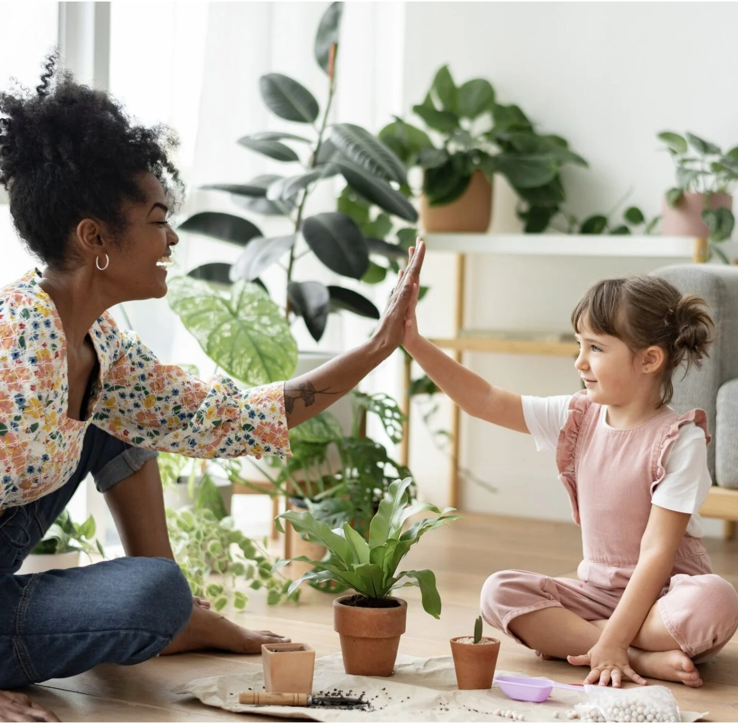 A woman and a young girl sitting on the floor, giving each other a high-five in a room filled with potted plants.