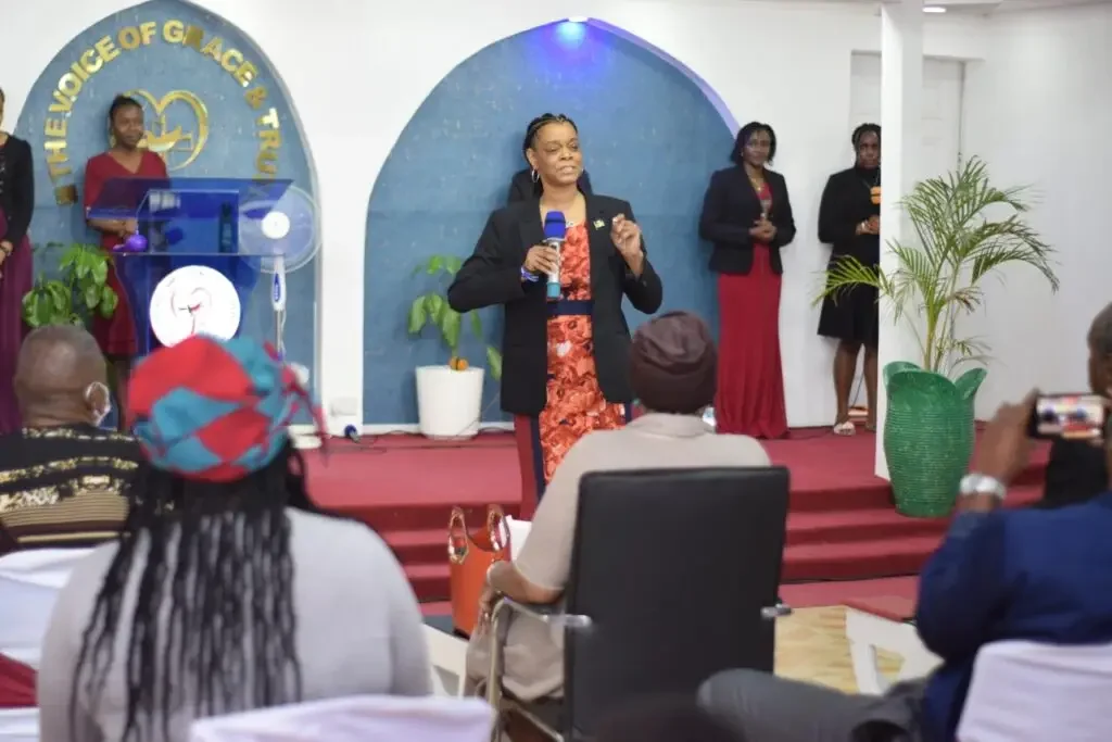 A woman in a black blazer and floral dress is speaking into a microphone on a stage with a blue and white background. Four women are standing behind her, and an audience is seated in front, with some taking photos.