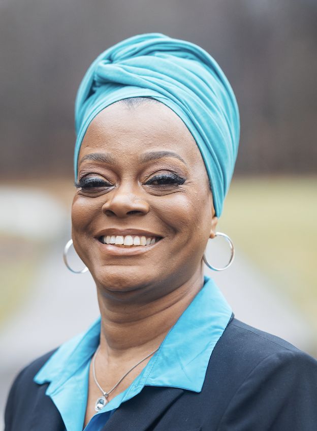 Portrait of a smiling woman wearing a blue headwrap, hoop earrings, and a black blazer with a blue shirt outdoors.