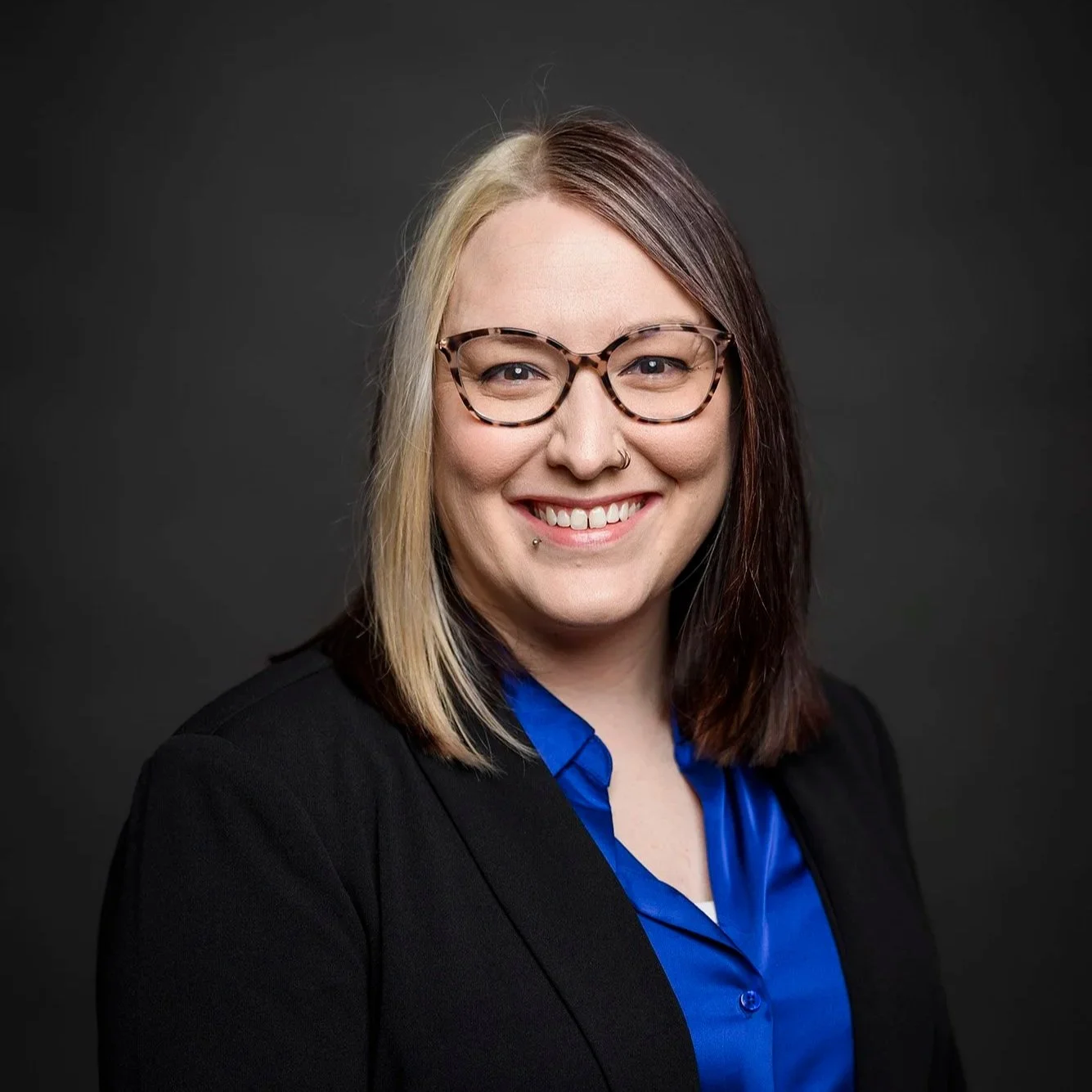 A headshot of Katie Maher, a white woman with half blonde, half dark brown hair wearing tortoise shell glasses, a black blazer with a royal blue silk shirt underneath on a dark gray background.