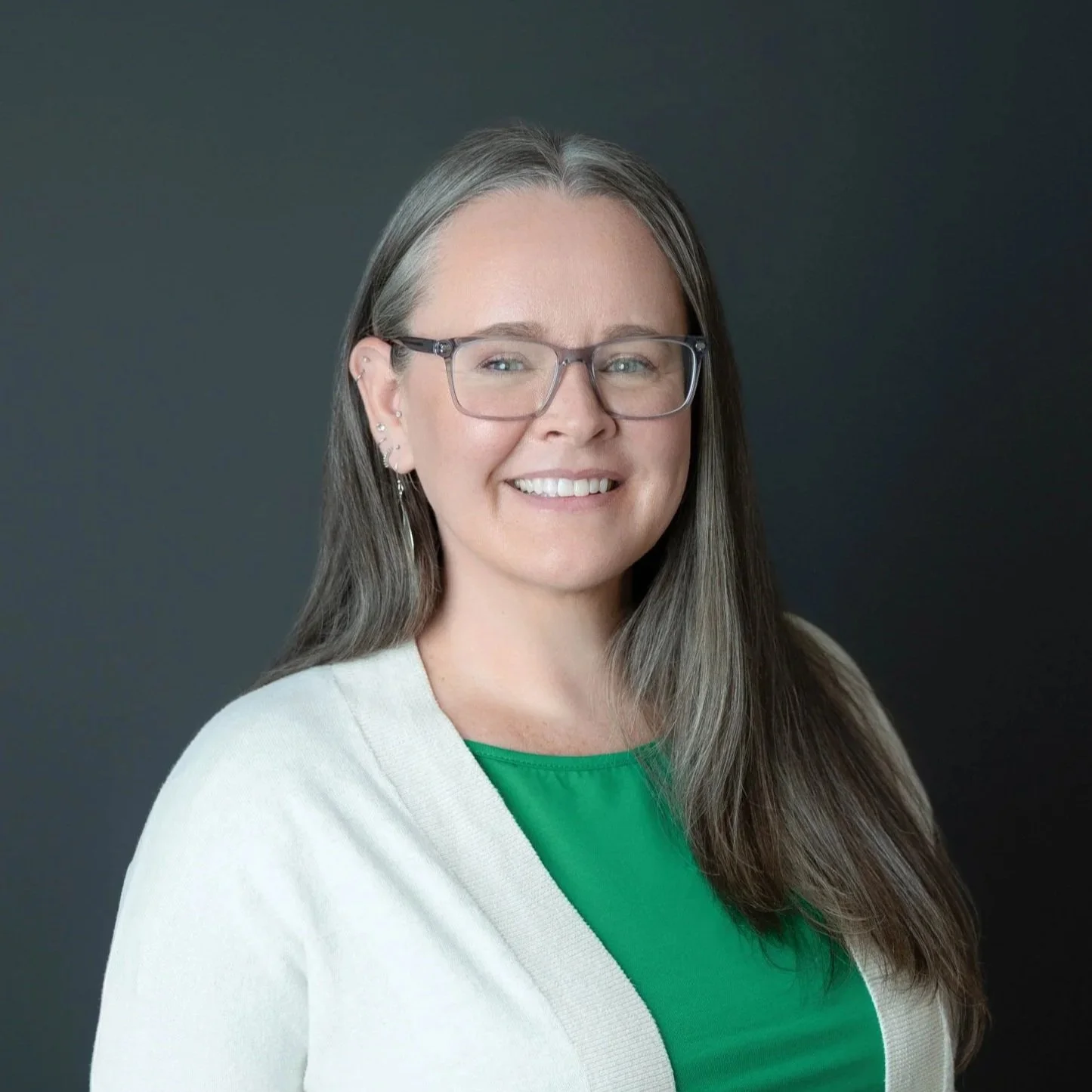 A headshot of Andrea Olsen, a white woman with gray square framed gloasses, long silver hair wearing a white sweater over a green shirt on a dark gray background.