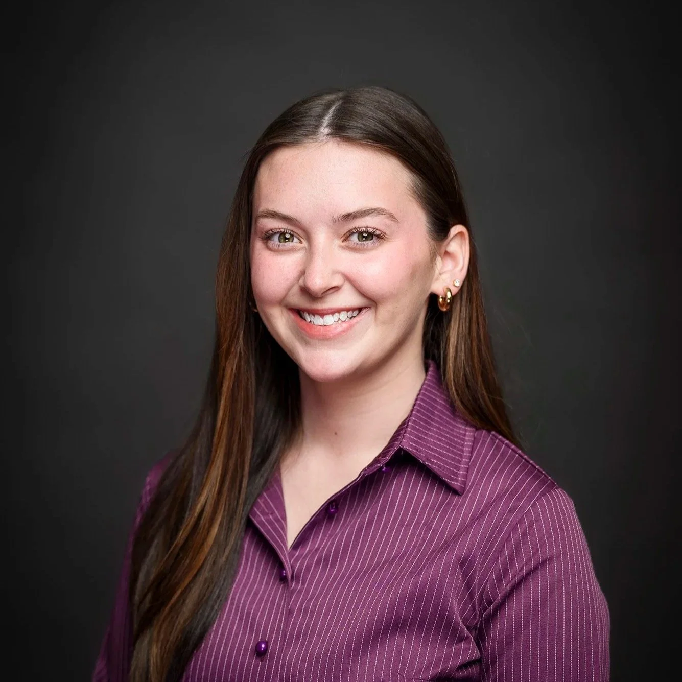 A headshot of Lily Shafer, a white woman with long dark brown hair wearing a striped purple button up on a dark gray background.