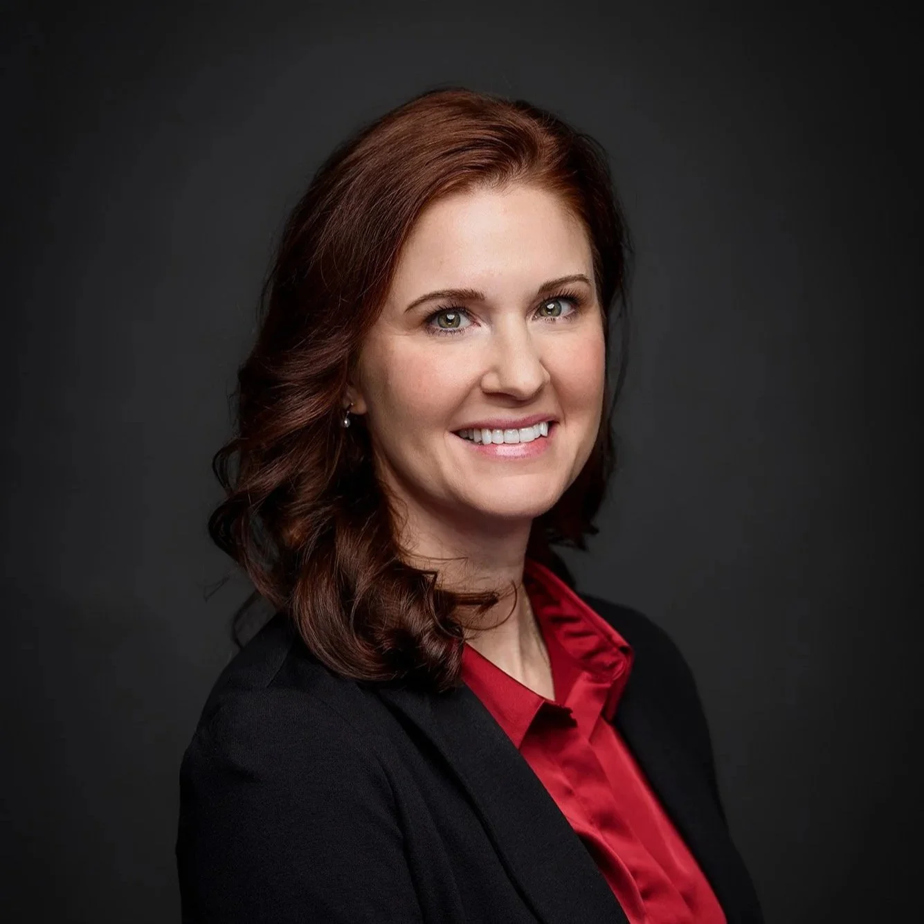 A headshot of Kaitlin Krueger, a white woman with dark auburn hair wearing a black blazer with a red silk button up underneath on a dark gray background.