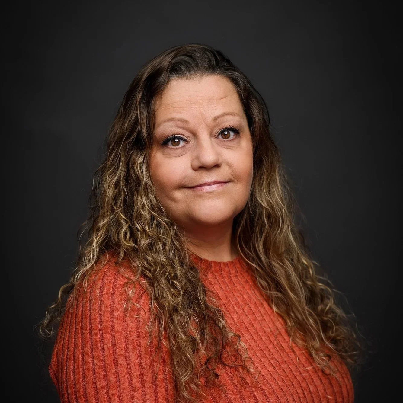 A headshot of Stephanie Schultz, a white woman with long brown curly hair wearing a red-orange ribbed sweater on a dark gray background.