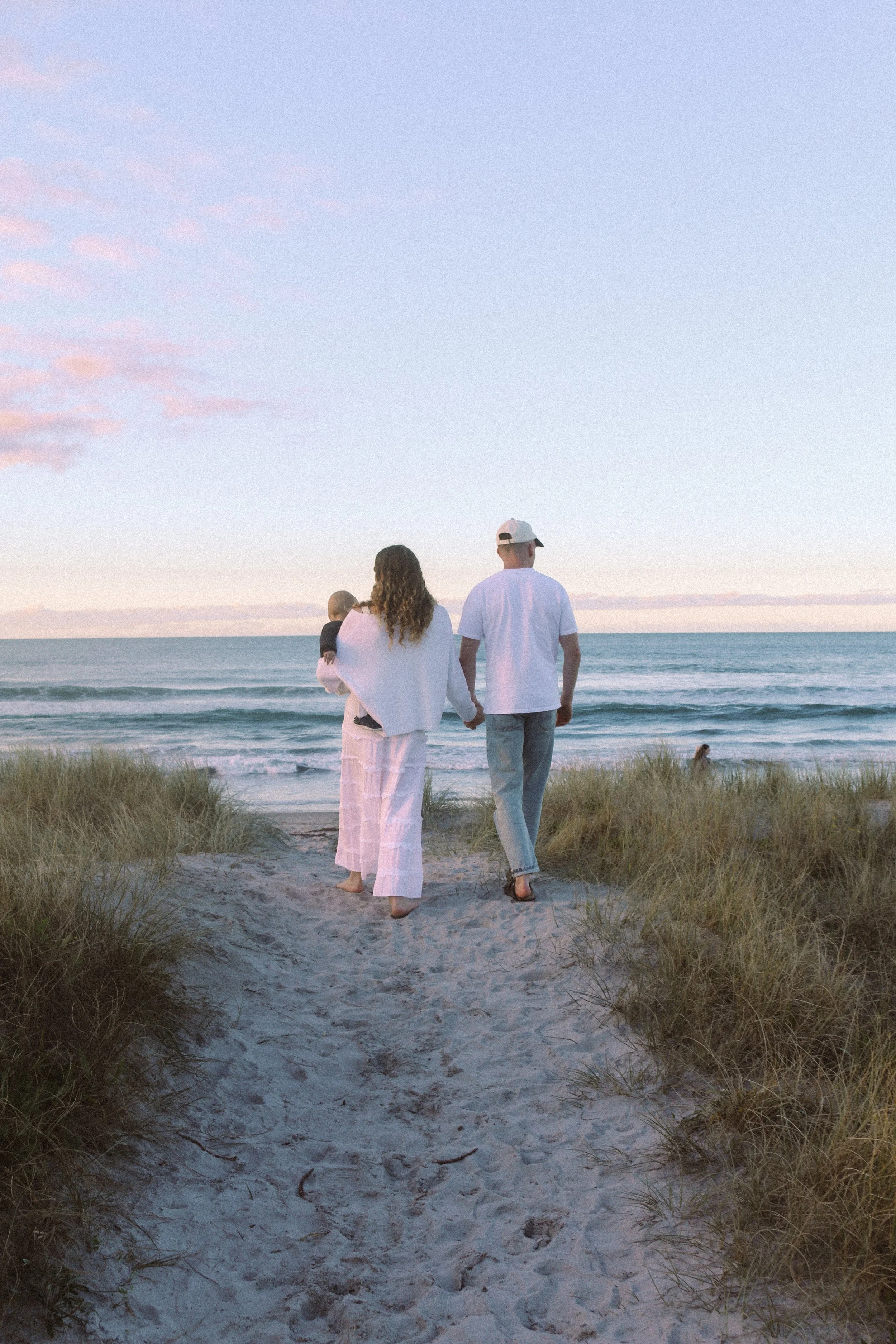 A family of three walking on a sandy beach at sunset, with tall grass in the foreground and the ocean in the background.