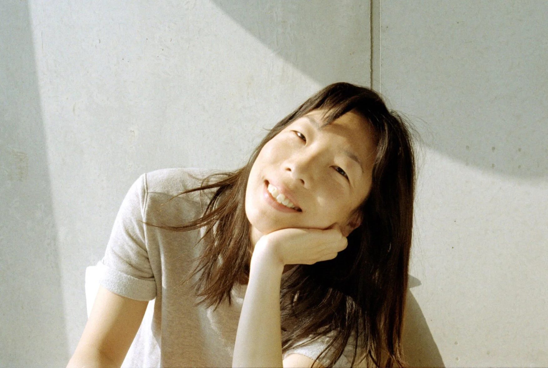 A young woman with long brown hair smiling and resting her chin on her hand against a plain light-colored wall.