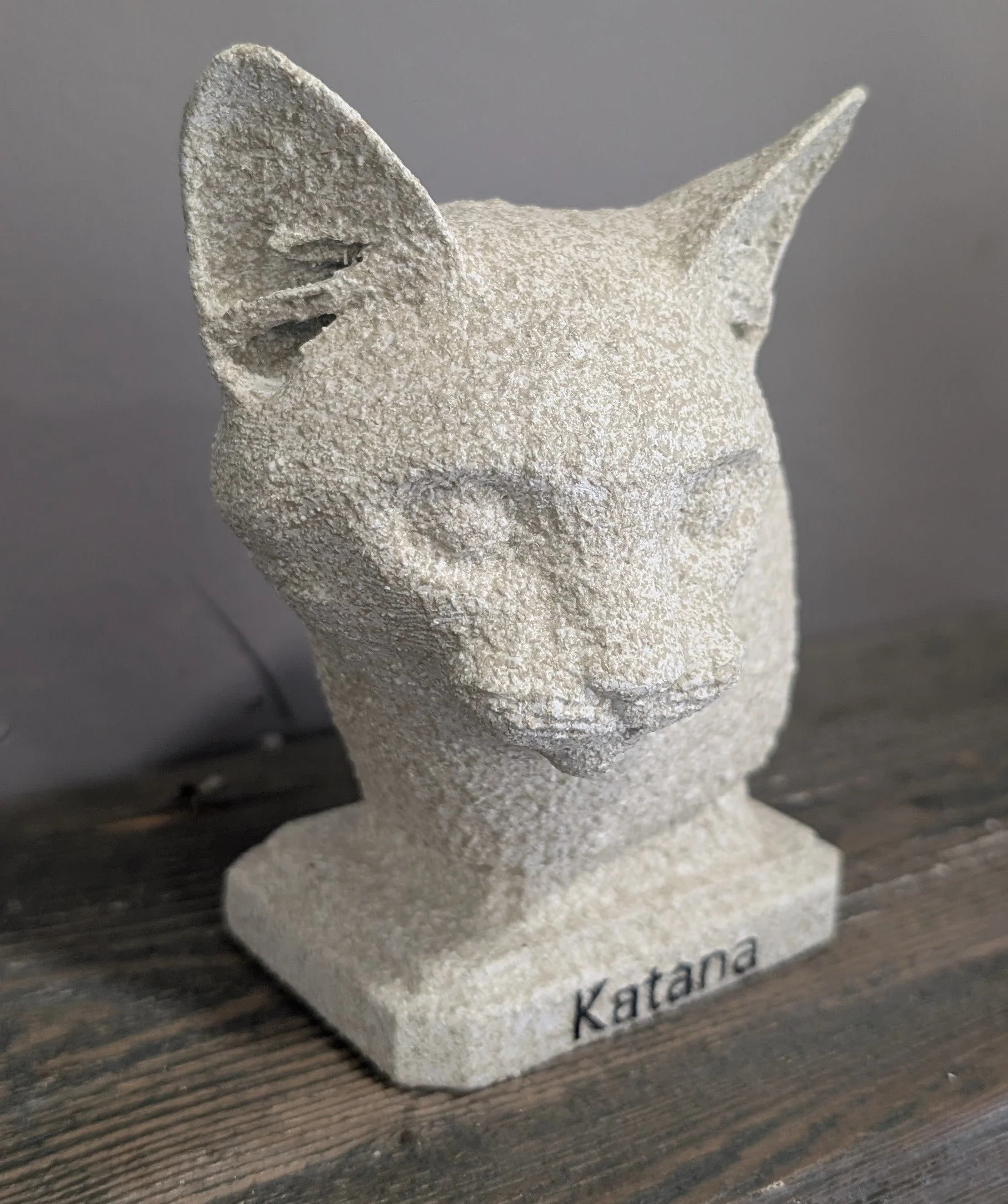 A sculpture of a cat's head made of textured stone, mounted on a base with the name 'Katana' engraved on it.