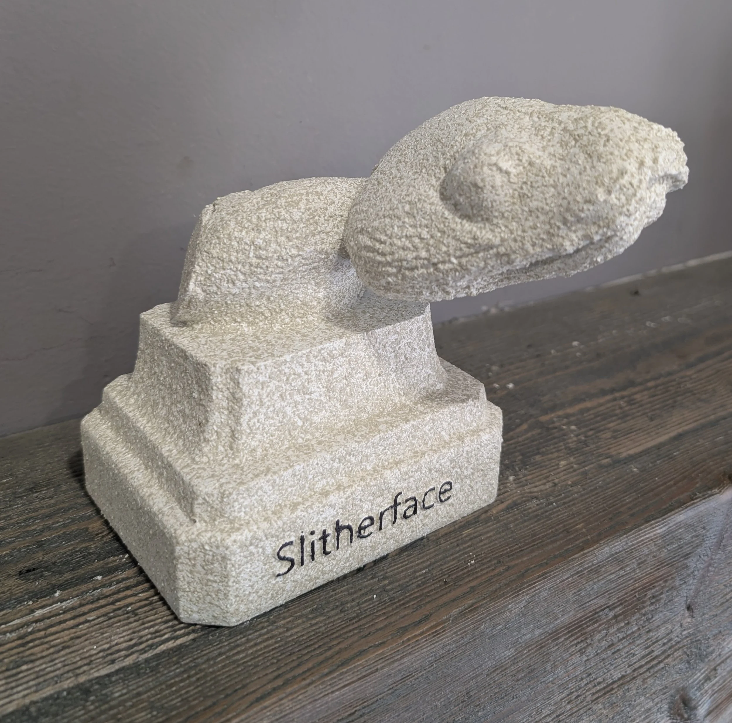 A stone sculpture of a snake with the word "Slithface" written on the base, placed on a wooden surface against a gray wall.