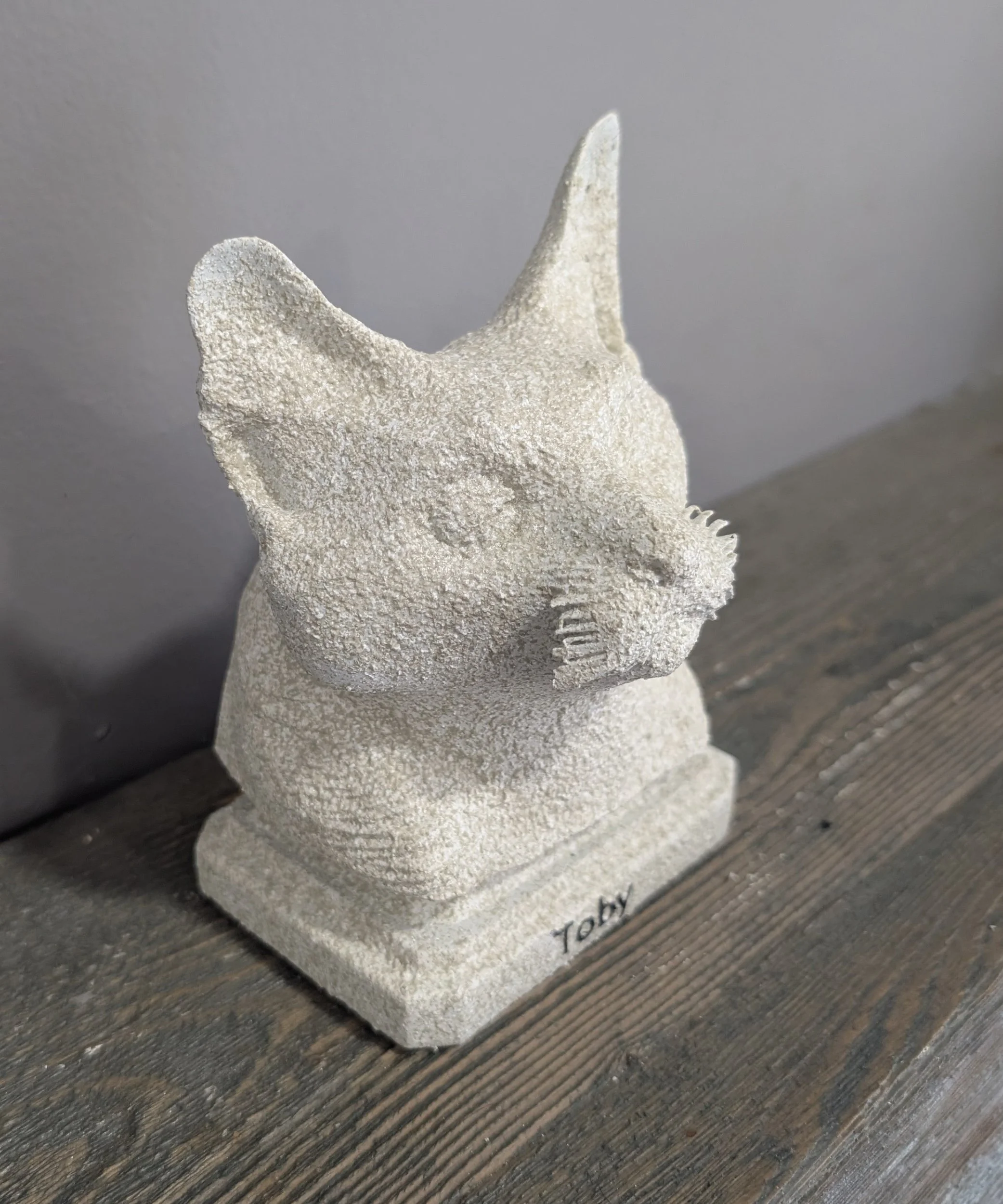 A textured sculpture of a cat's head with one ear pointed up and the other slightly back, mounted on a rectangular base with the word "Toby" inscribed on it, placed on a wooden surface against a gray wall.
