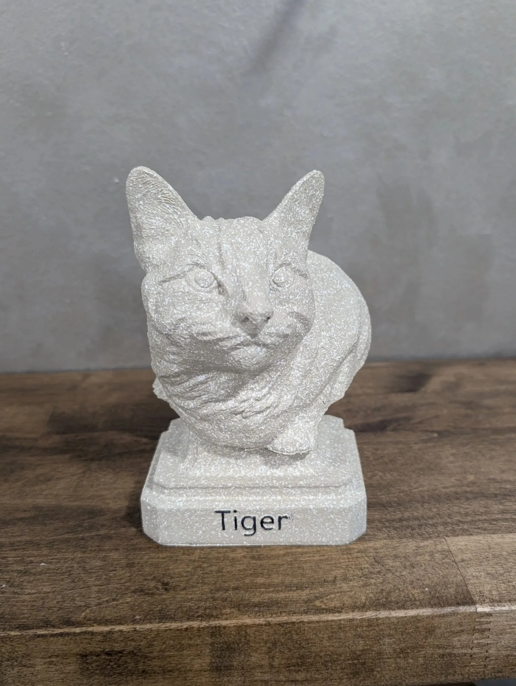 A stone cat figurine on a square base with the word 'Tiger' written on it, placed on a wooden surface against a grey background.
