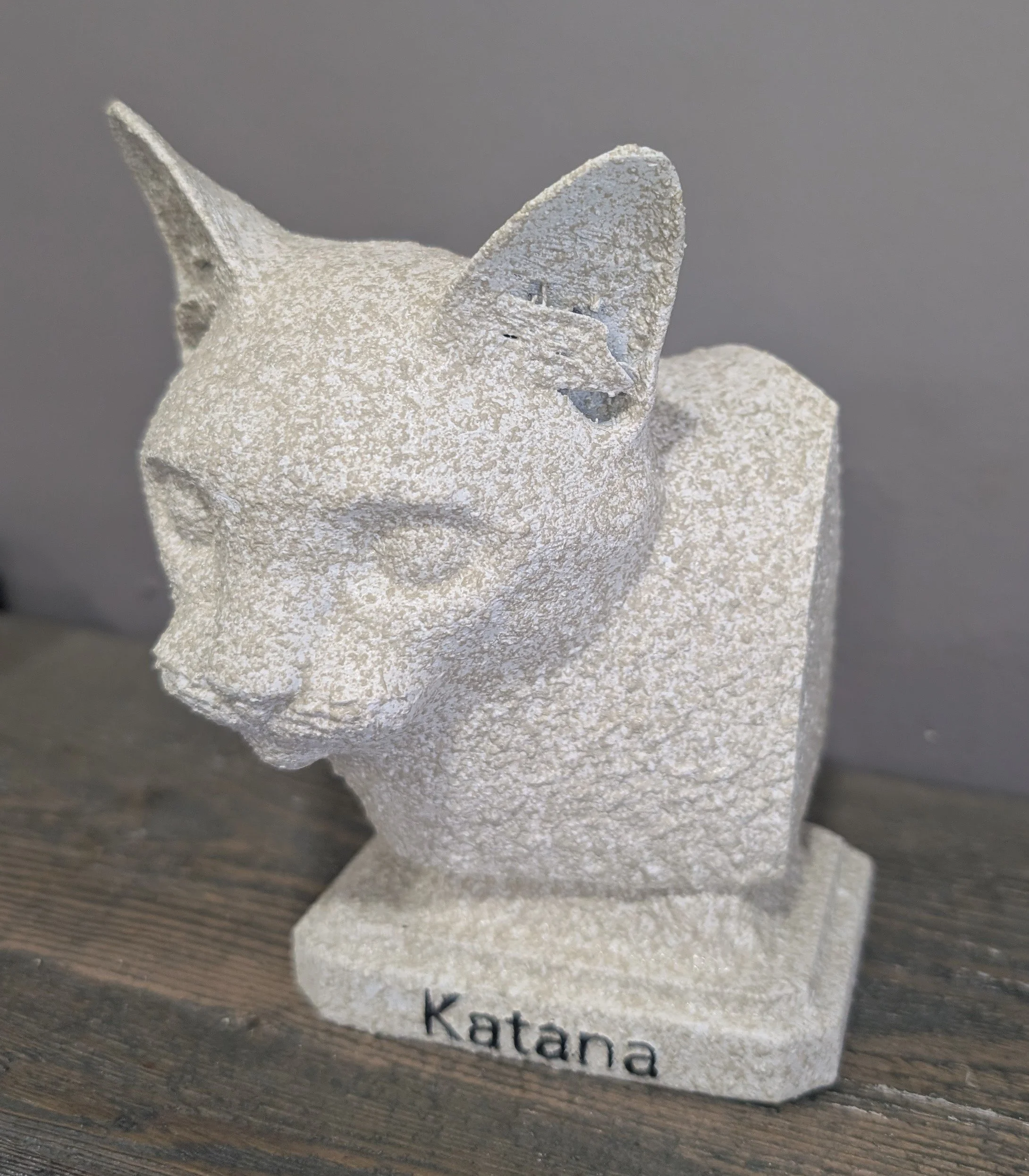 Stone sculpture of a cat's head with a small bird perched on its ear, on a rectangular base with the word 'Katana' engraved.