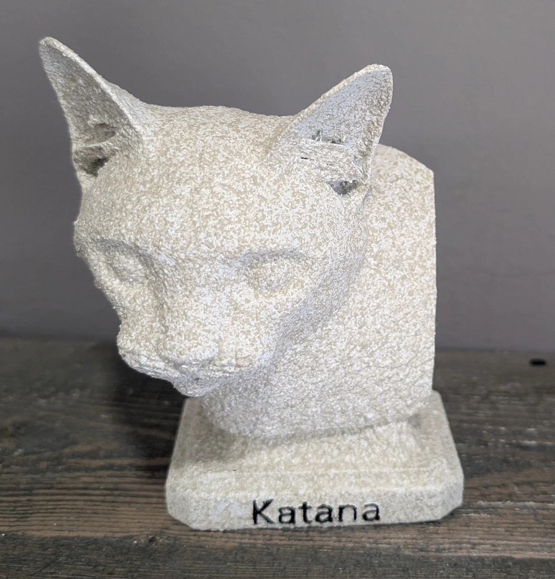 A stone sculpture of a cat's head on a square base with the word 'Katana' engraved at the front