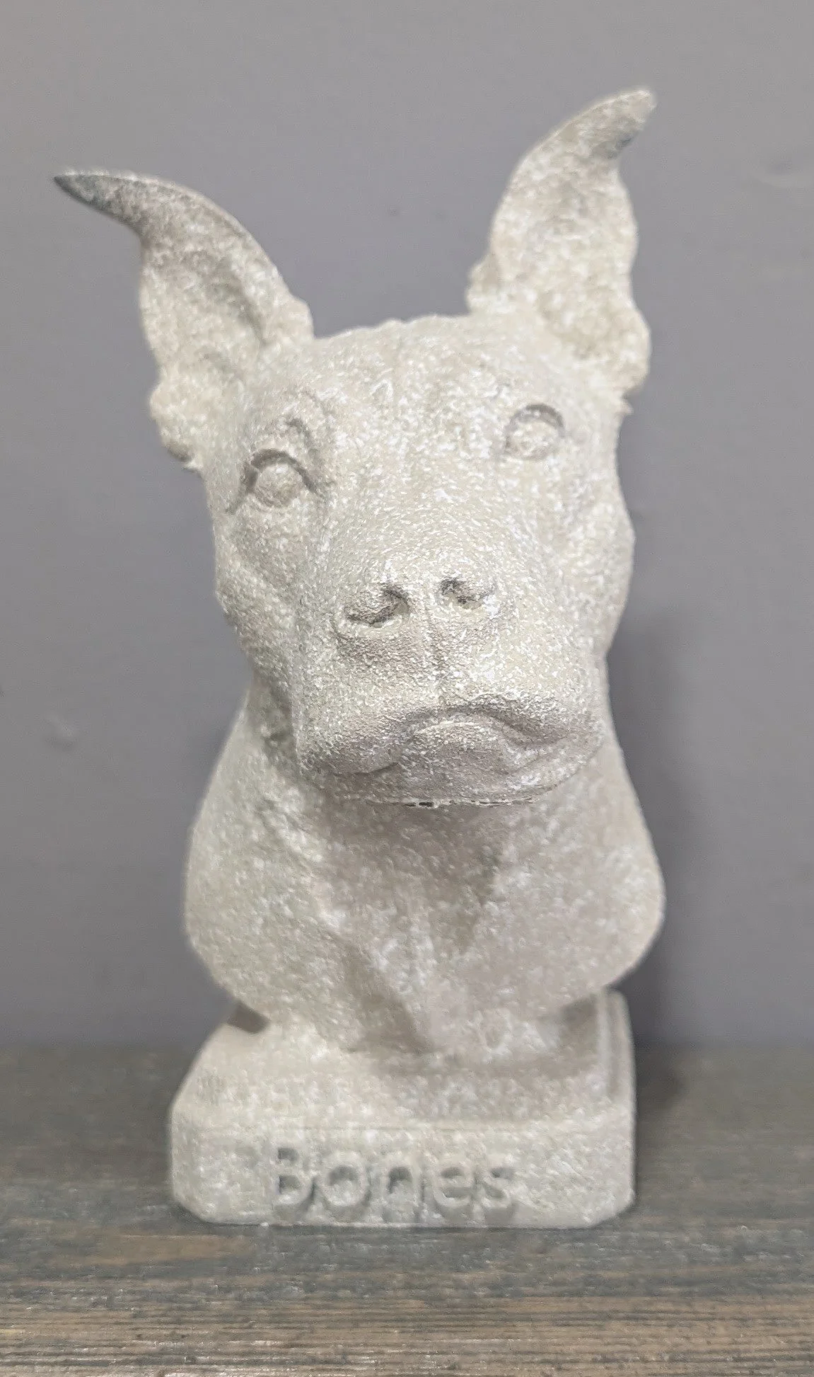 A carved stone sculpture of a dog with a textured surface, pointed ears, and a serious expression. The sculpture is mounted on a base with the word 'DOGS' engraved on it.