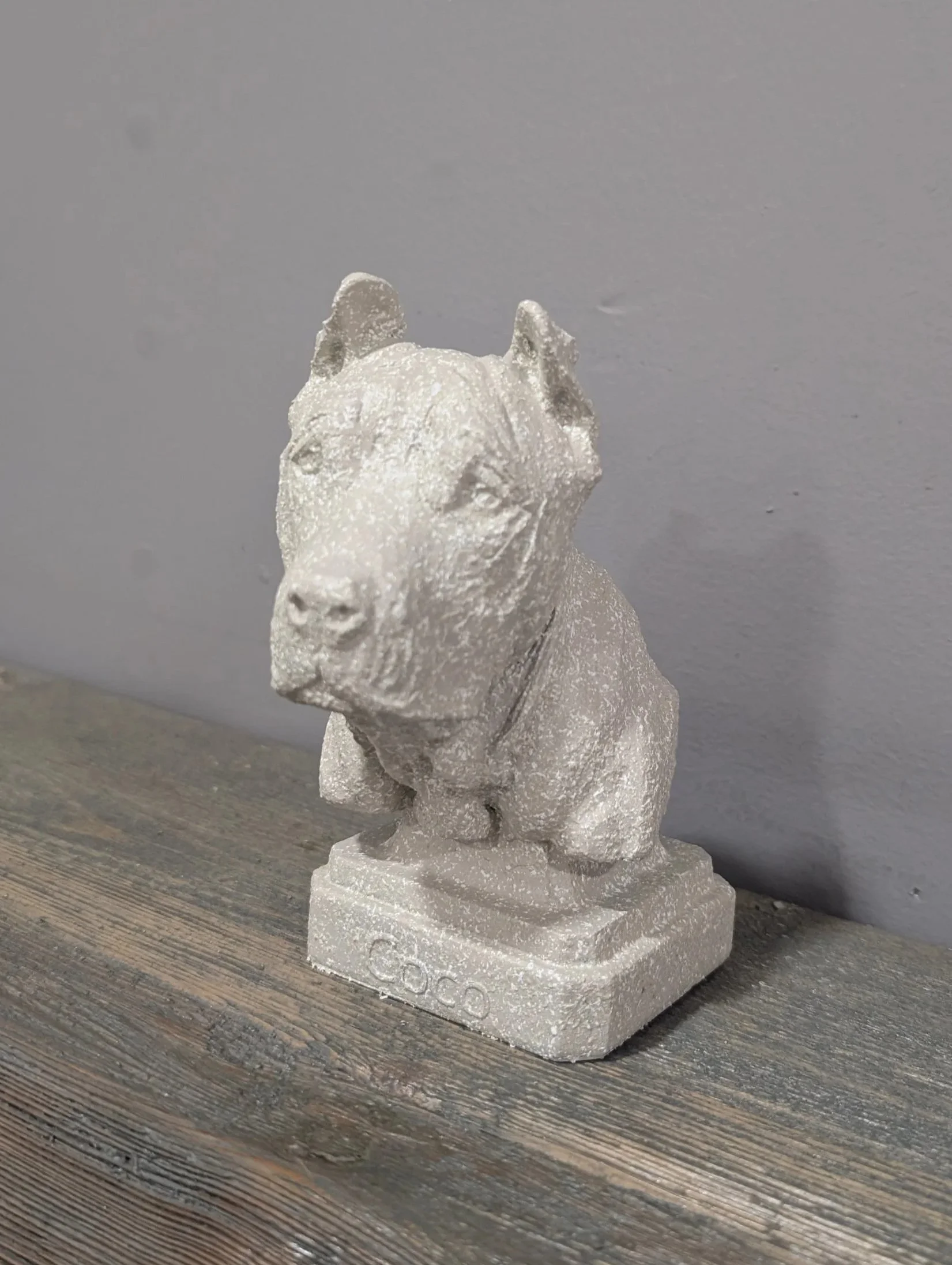 Small statue of a dog on a wooden surface against a gray wall, with the word 'COCO' inscribed on the base.