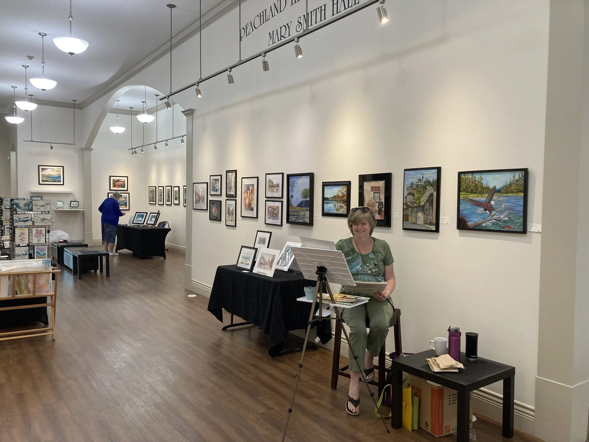 An art gallery with paintings on the wall, including landscapes and cityscapes. A woman is sitting at a small table with art supplies, smiling, with framed artworks displayed nearby. Two people are viewing art in the background, and the gallery has a wooden floor and white walls.