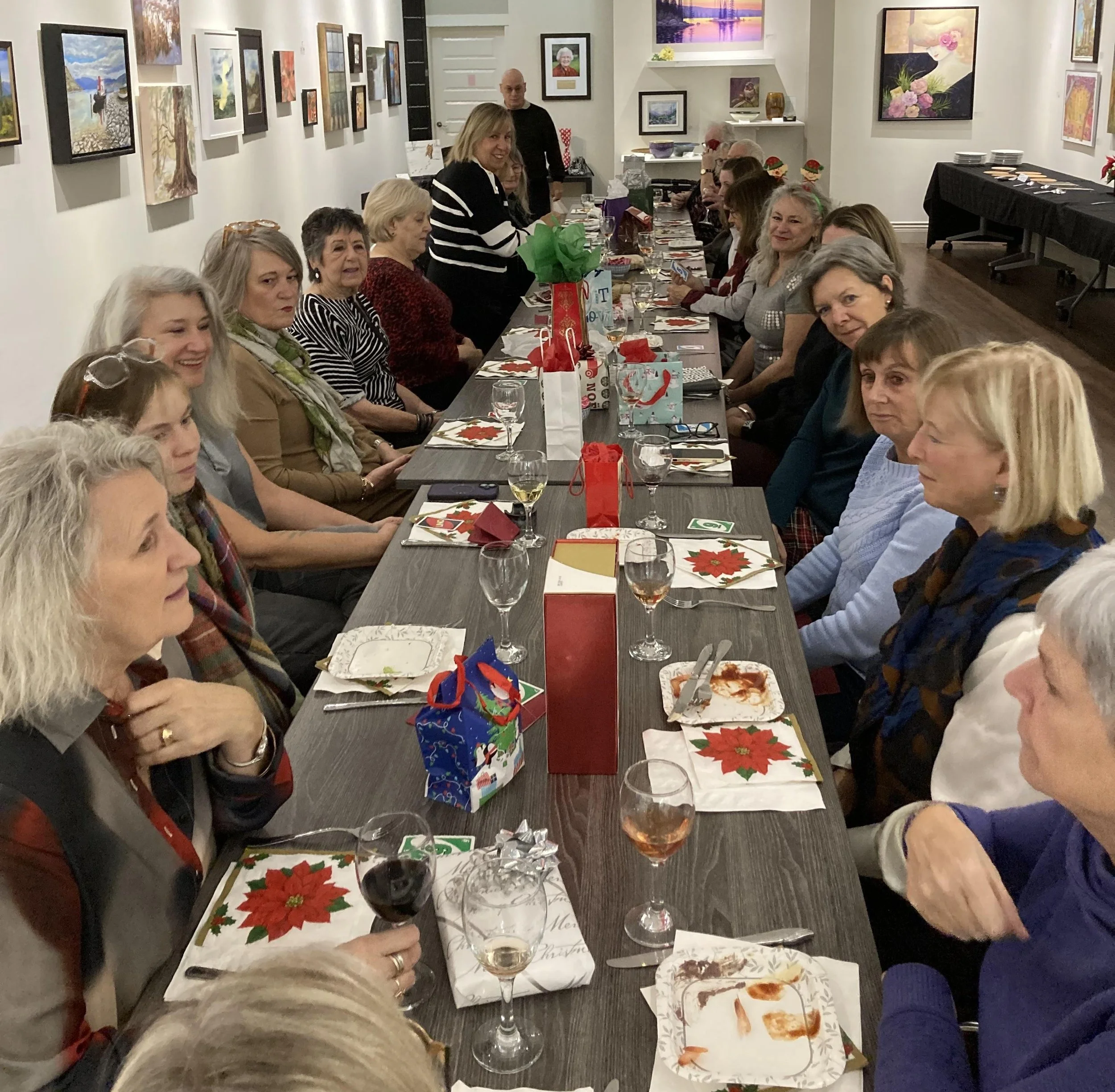 People gathered around a long table for a holiday celebration, with Christmas-themed decorations and gifts, in an art gallery or similar venue with paintings on the walls.