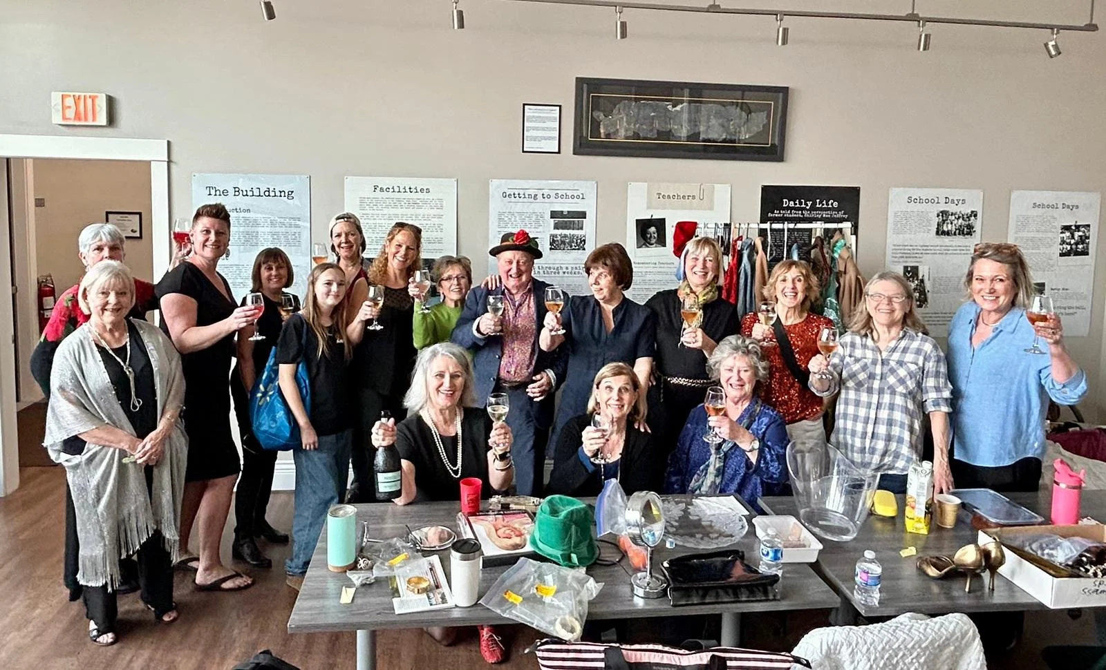 A large group of people, mostly women, celebrating together in a room with historical posters on the wall, holding glasses of wine or champagne, some wearing festive hats and colorful attire, with a table in front of them filled with various items including bottles, cups, and decorations.