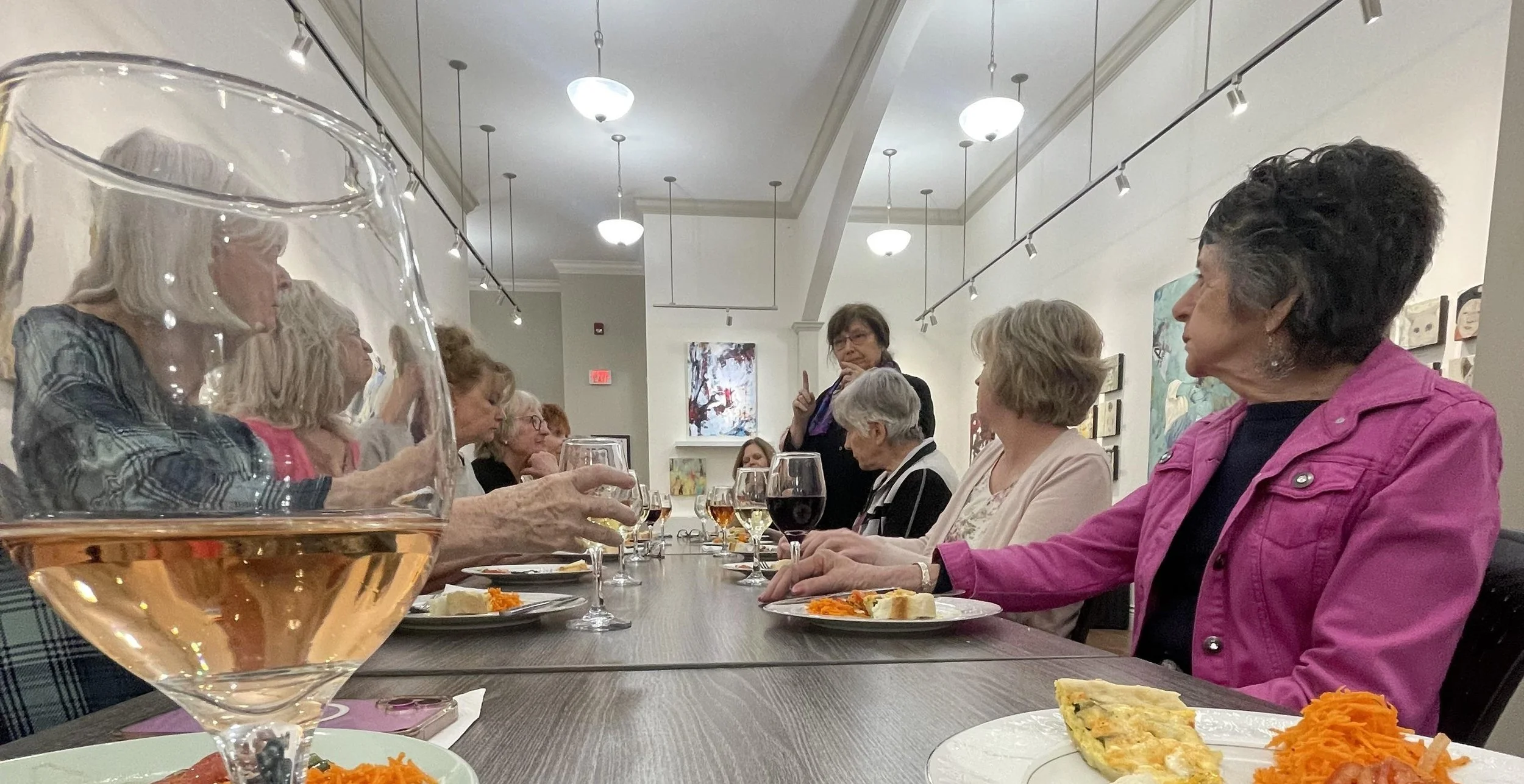 A group of older women sitting at a long table in an art gallery, having a meal and drink, with a woman standing and talking to them. There are glasses of wine and plates of food on the table.