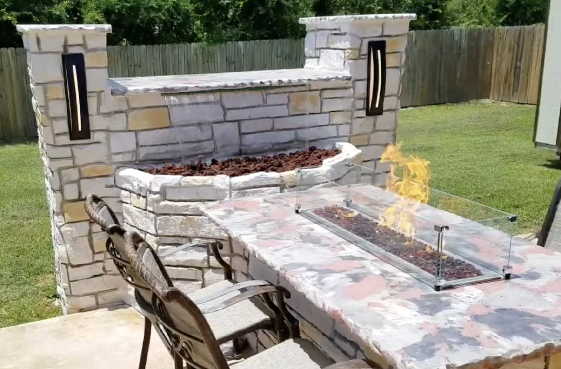 Outdoor backyard with stone fireplace and built-in fire pit, with a glass guard, surrounded by lawn and fence.