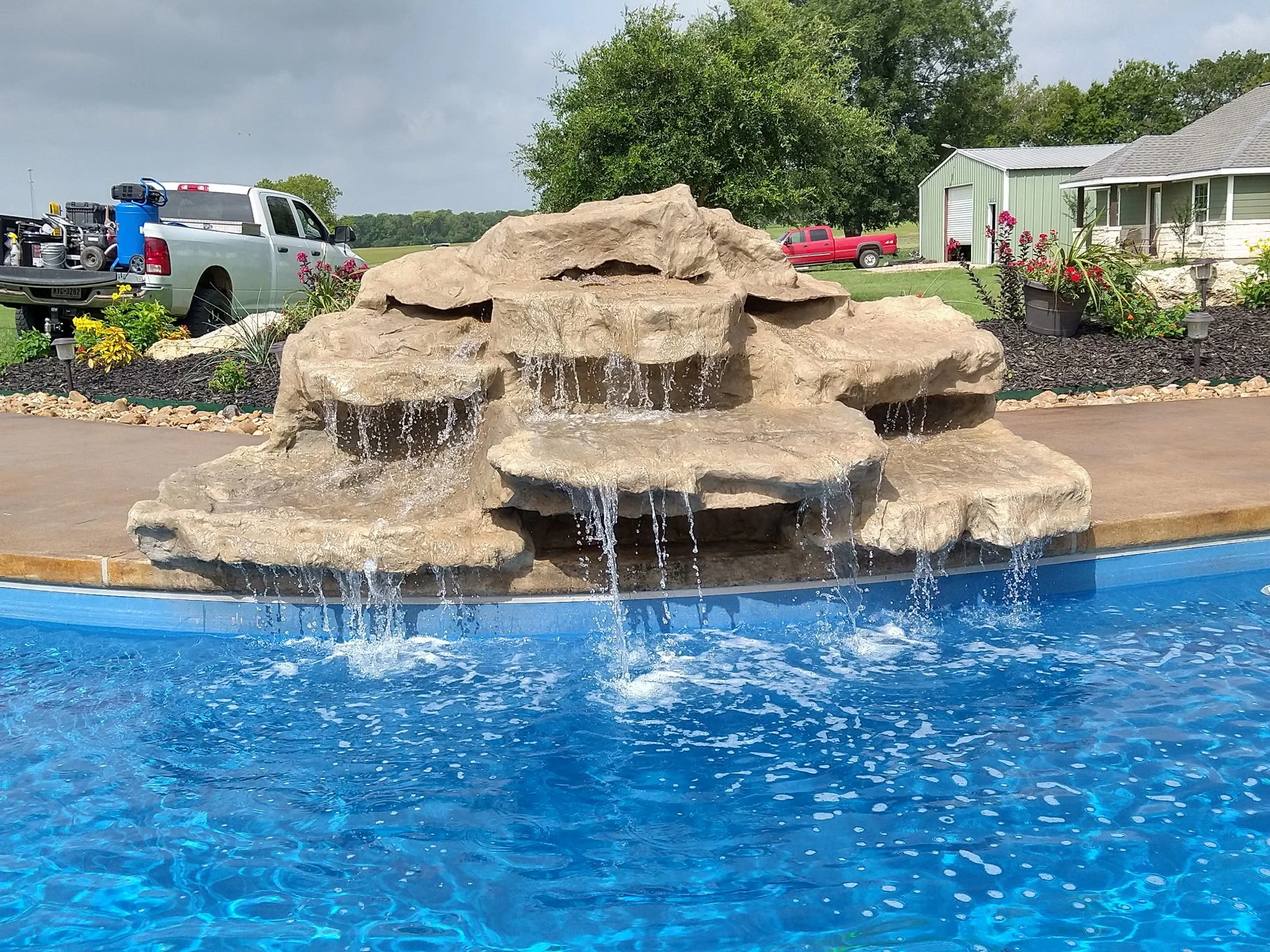 A backyard with a swimming pool featuring a rock waterfall. There are trucks and a house with a garden in the background.