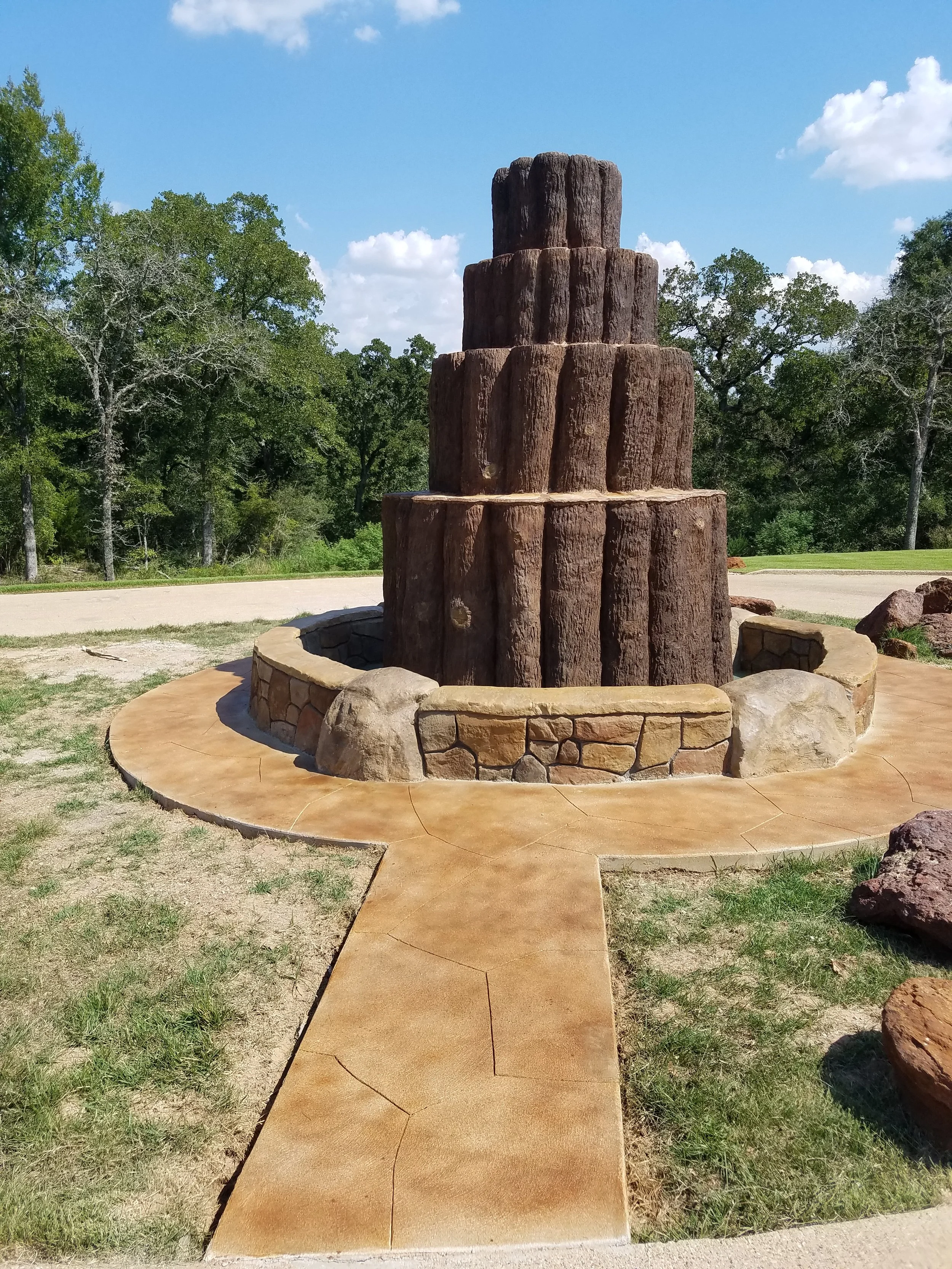 A multi-tiered stone and wood fountain in a park with trees and blue sky in the background.