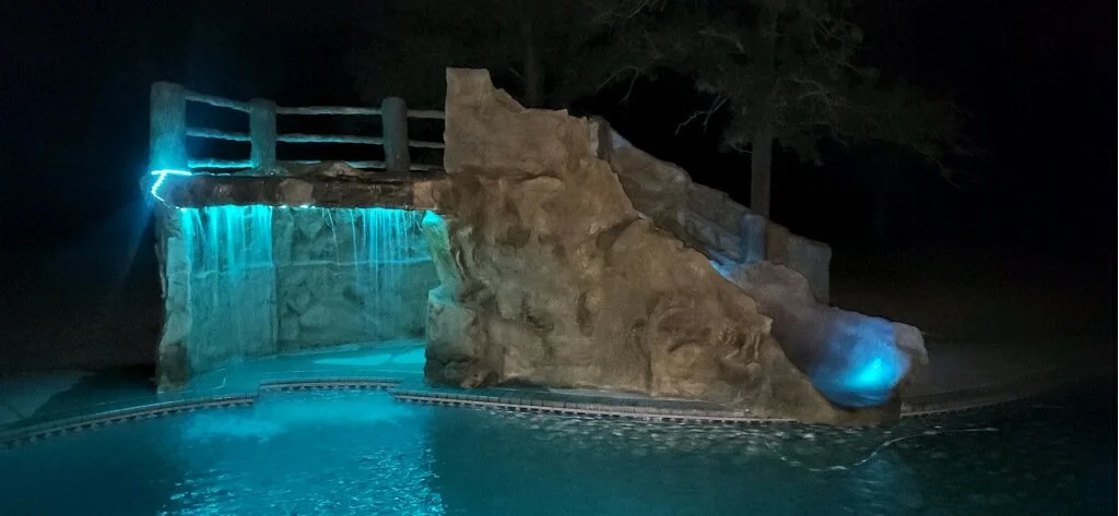 Nighttime scene of an outdoor pool with illuminated waterfalls and rocks, glowing blue lights, and slide at the top.