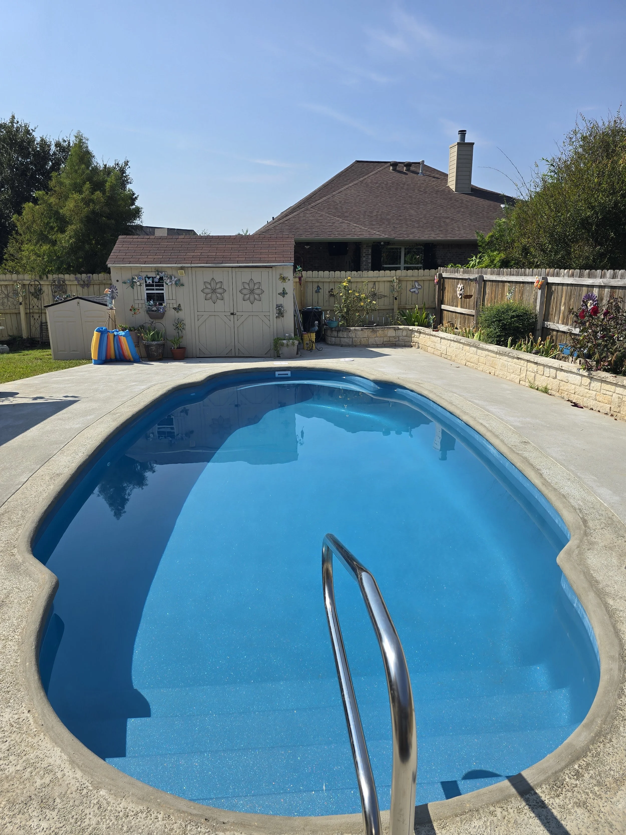 Backyard with an in-ground swimming pool, small shed with flower decorations, wooden fence, and a brick house in the background, under clear blue sky.