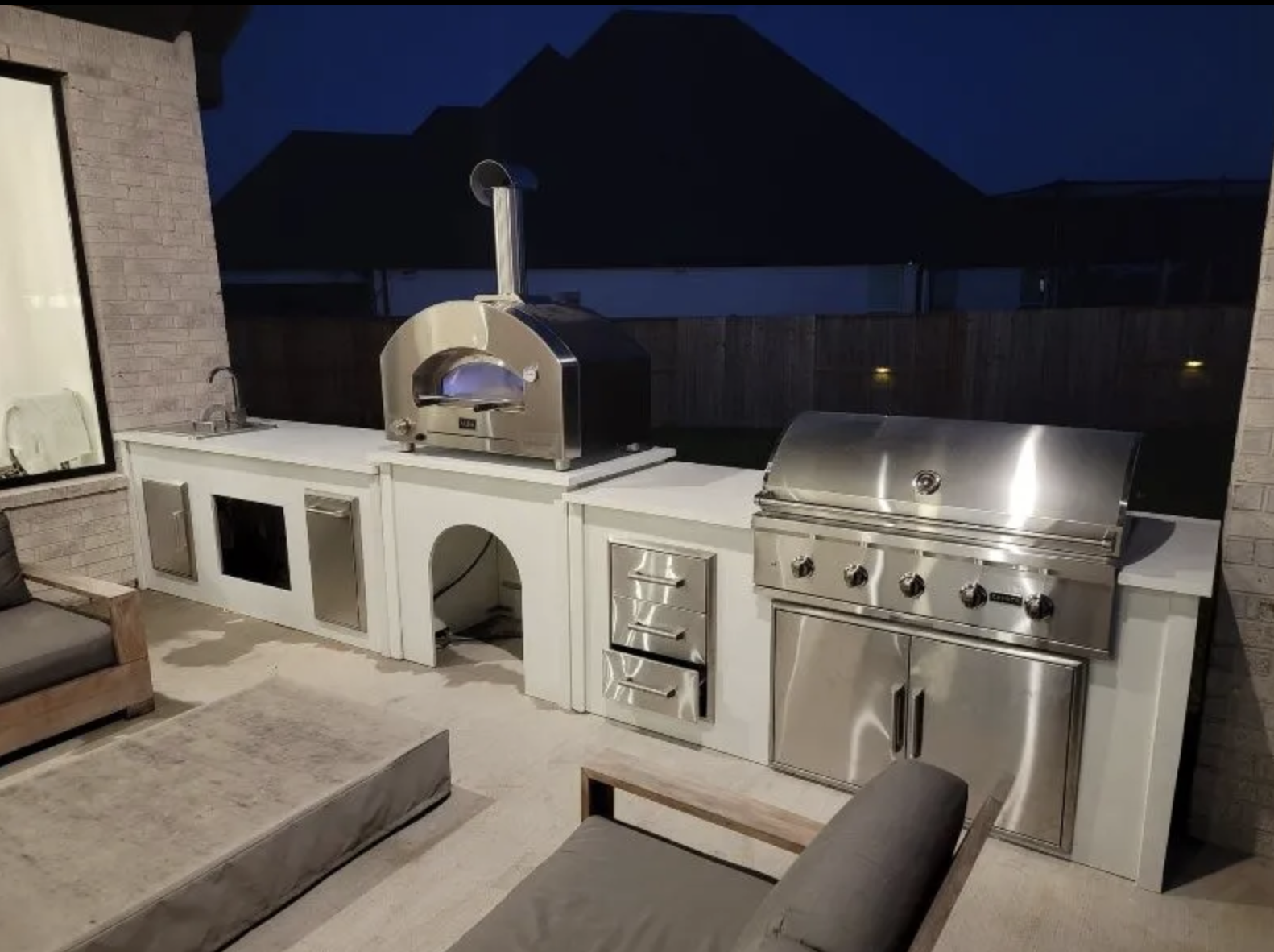 Outdoor kitchen with stainless steel grill and pizza oven under dark evening sky.