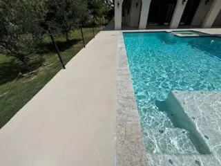 Part of a swimming pool with clear blue water and a concrete deck outside. The pool has built-in steps and is adjacent to a grassy yard with trees.