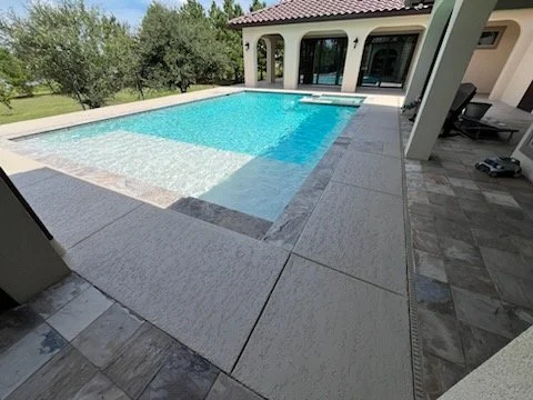 Backyard with a large in-ground swimming pool surrounded by a concrete patio and a house with arches in the background.