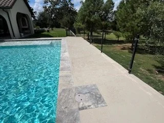 View of a backyard swimming pool with a concrete deck and black safety fencing, trees, and grassy area in the background.