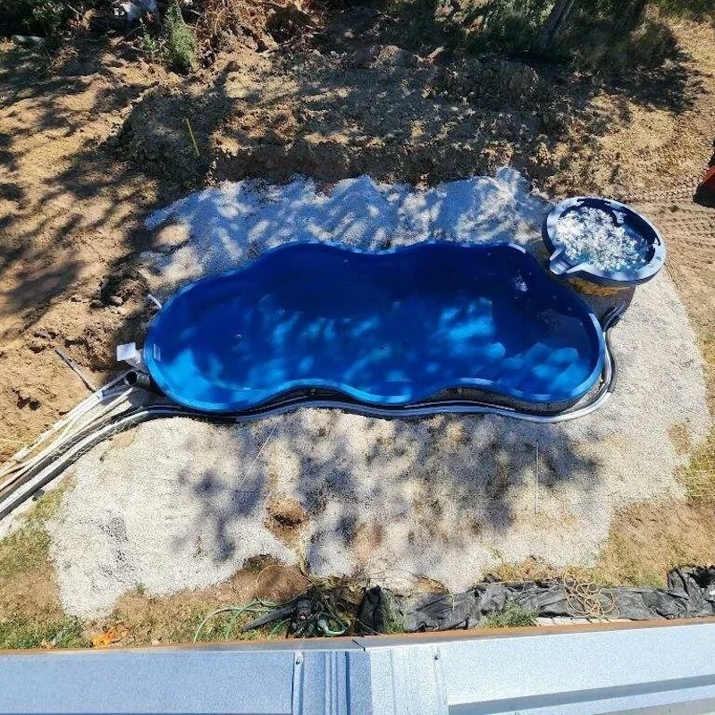 A small blue in-ground swimming pool filled with water on a concrete pad, with a bucket of water nearby, surrounded by dirt and some trees casting shadows.