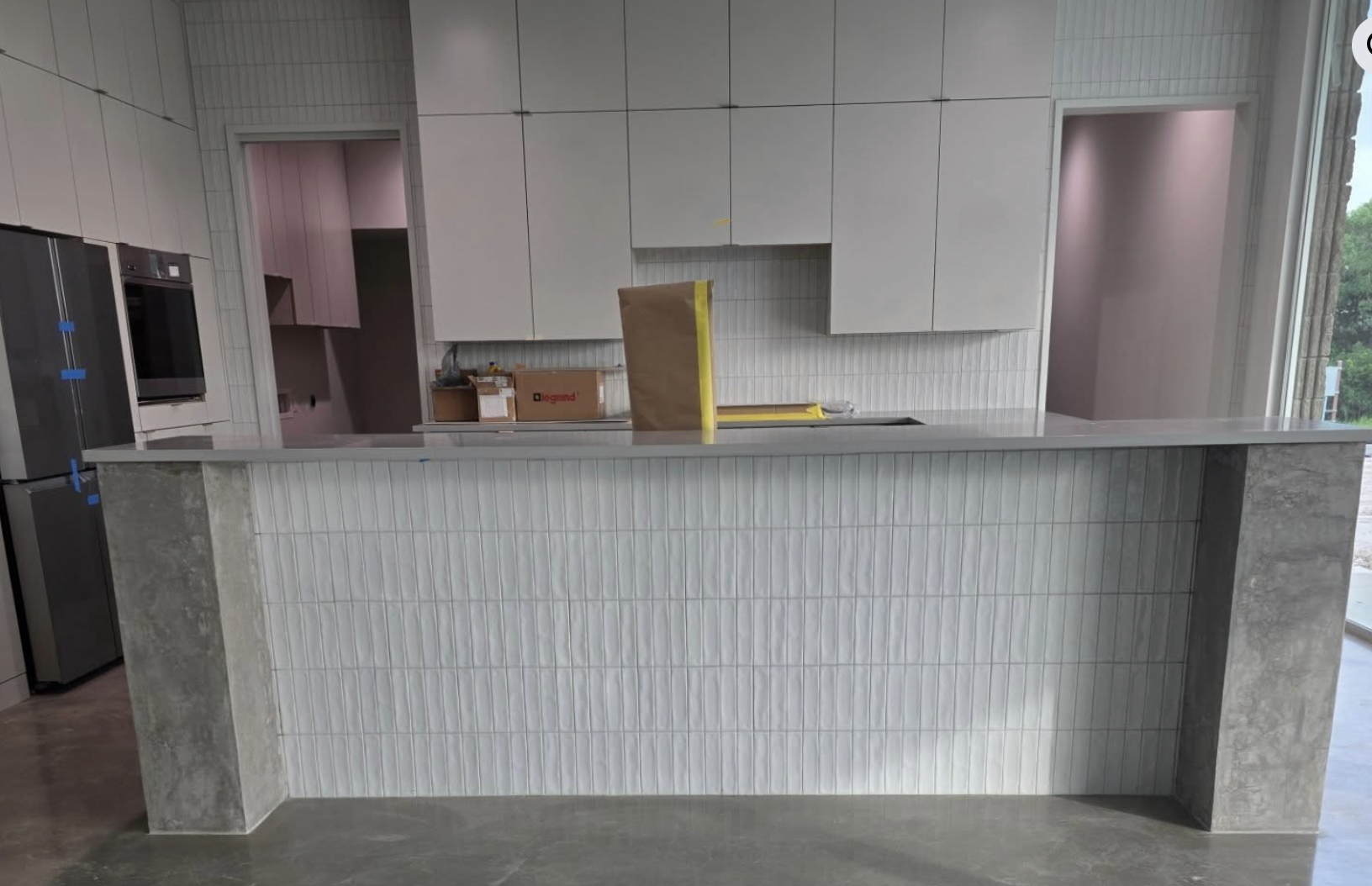 Modern kitchen under construction with white cabinetry, gray concrete countertops, and a large island with a textured white front and concrete sides. A window on the right shows greenery outside.