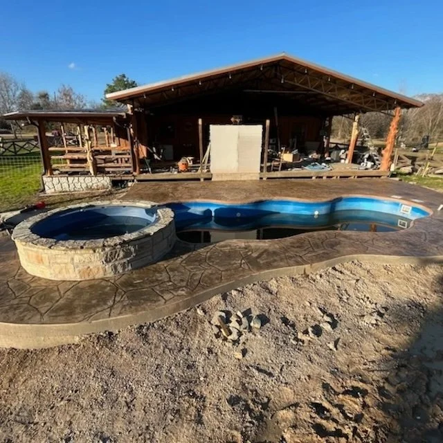 Outdoor area with an in progress pool and hot tub in front of a wooden pavilion.