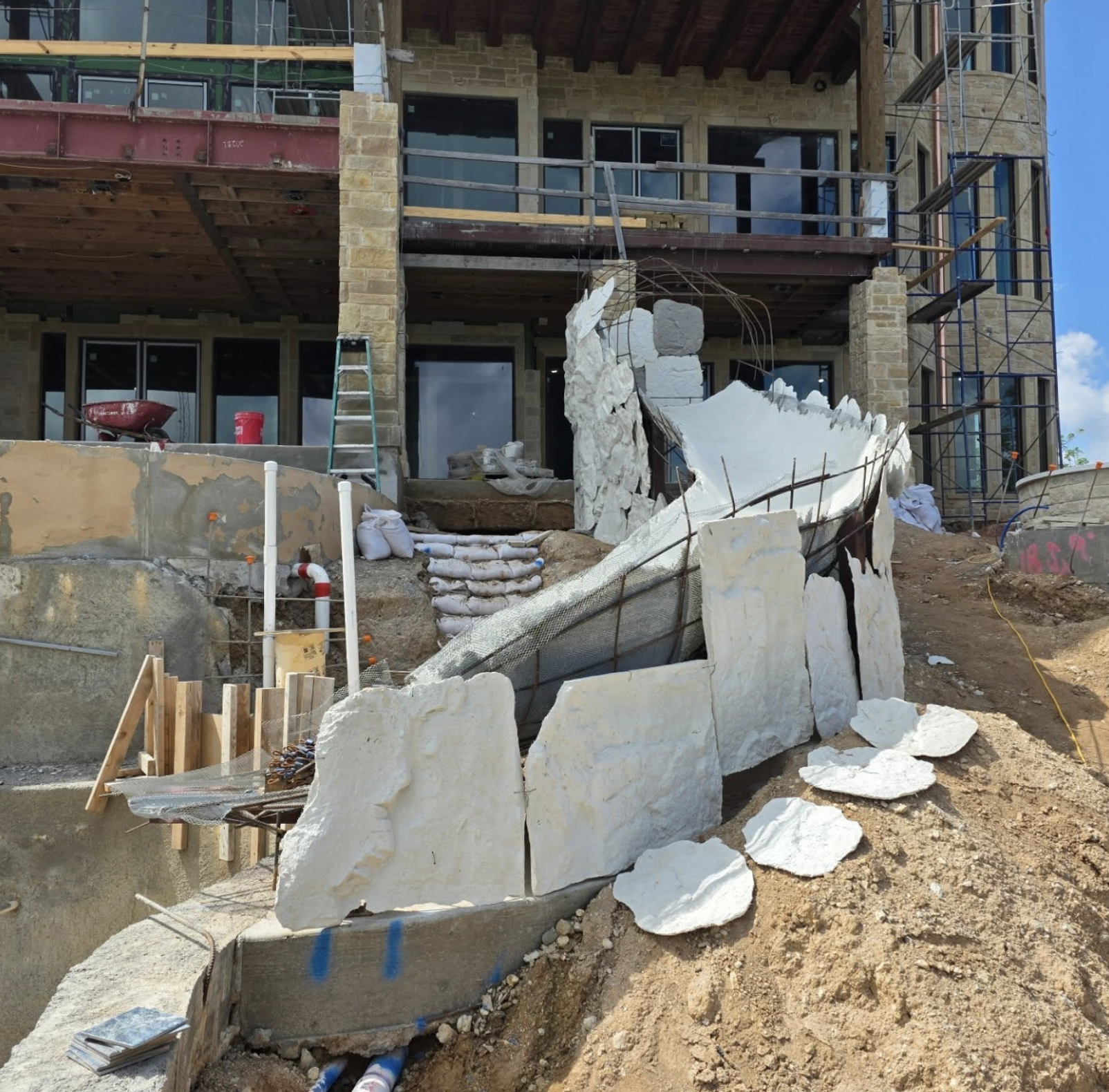 Construction site of a multi-story building with scaffolding, a ladder, and large rocks being placed as part of the retaining wall or landscaping feature in front of the building.