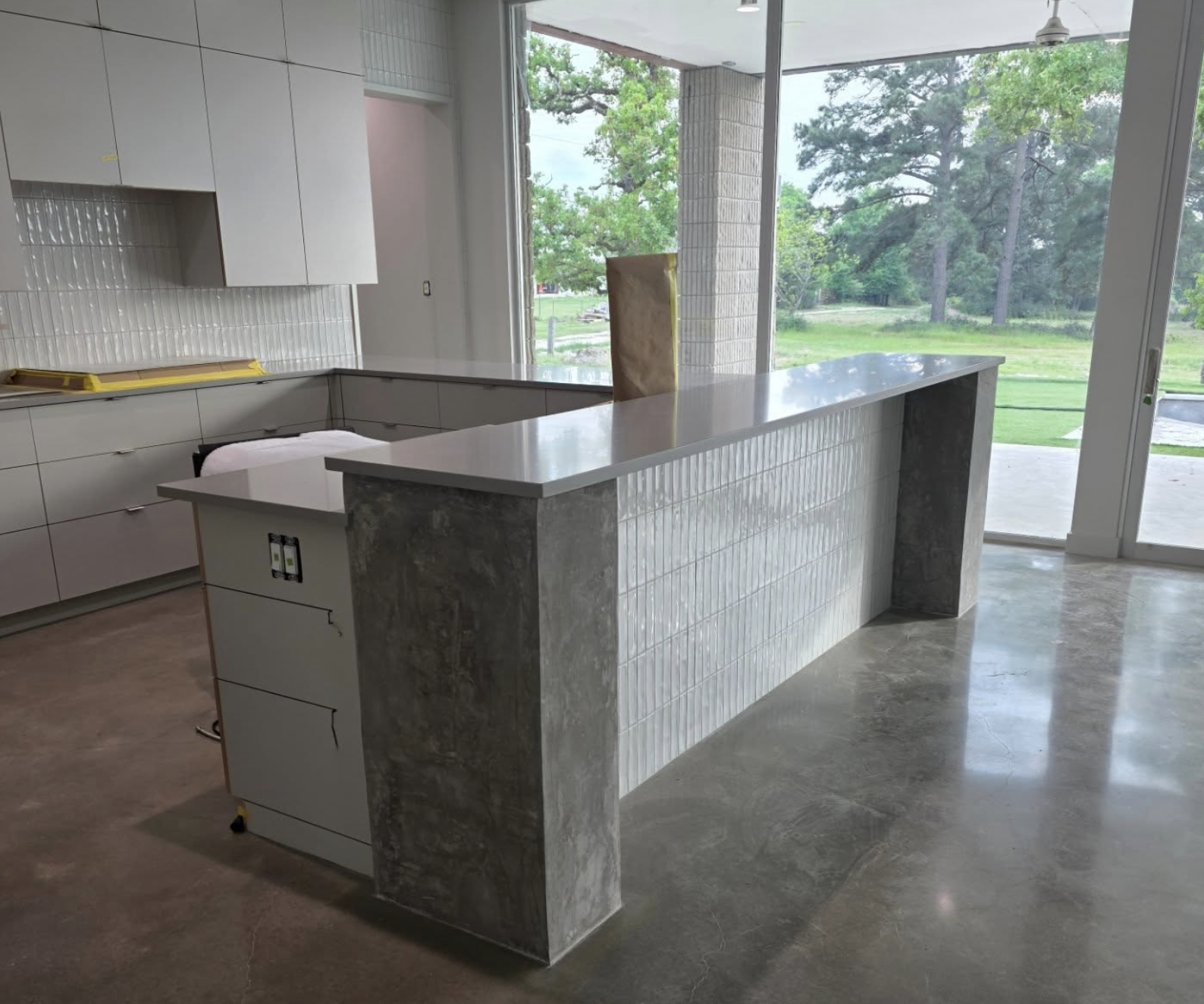 Modern kitchen with a large white island featuring a gray stone accent on the sides, white cabinetry, and large windows overlooking a green outdoor area.