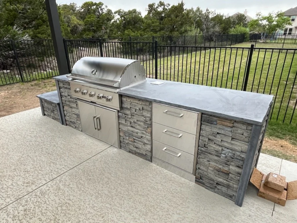 Outdoor grilling station with a built-in stainless steel grill, stone facade, and a concrete countertop, situated on a patio with a black metal fence and a grassy yard in the background.