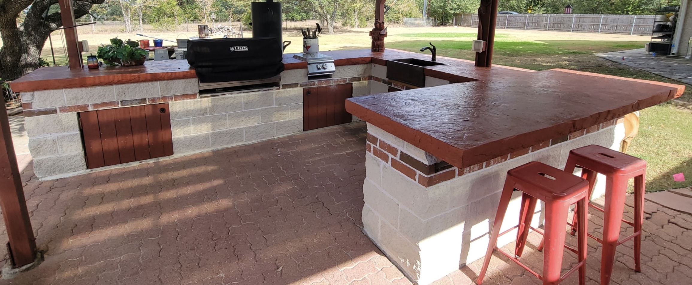 Outdoor kitchen with a brick and stone counter, black grill, small water basin, and red stools, set on a paved patio area with a grassy yard in the background.