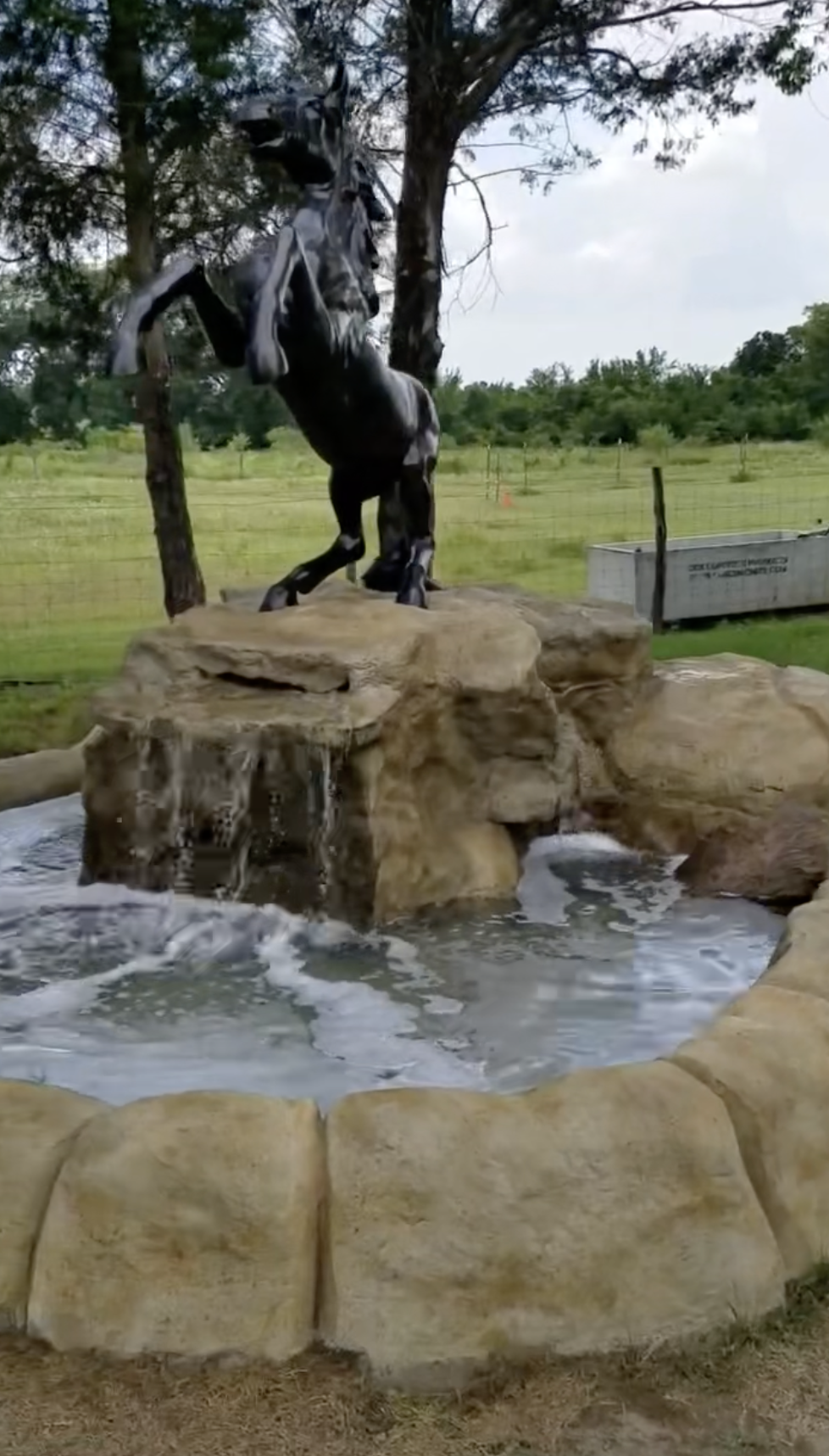 A black horse statue on a rock formation with a small waterfall and pool in front, surrounded by grass and trees.