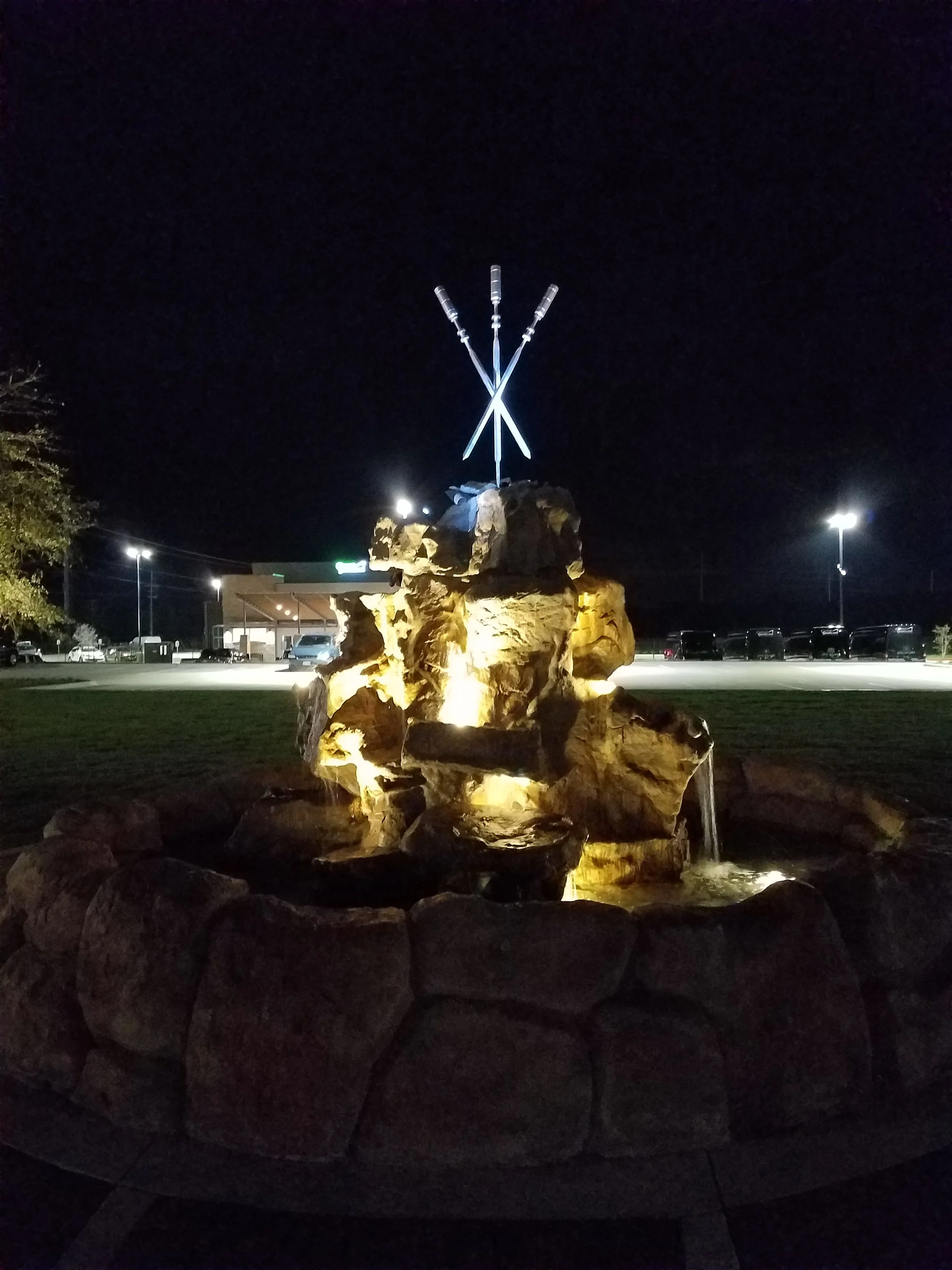 Night view of a lit rock fountain with three illuminated spears or lances crossing above it, located in a parking lot with streetlights and a building in the background.