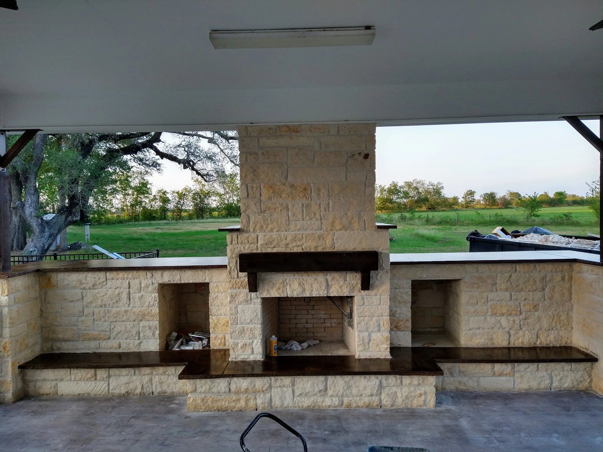 Exterior view of a stone fireplace with wooden mantel on an outdoor patio, overlooking a green lawn and trees with a gray sky.