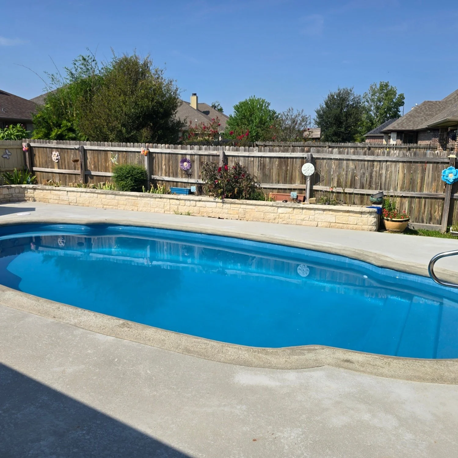 Residential backyard with a swimming pool, wooden fence decorated with colorful paper flowers, and lush trees in the background under a clear blue sky.