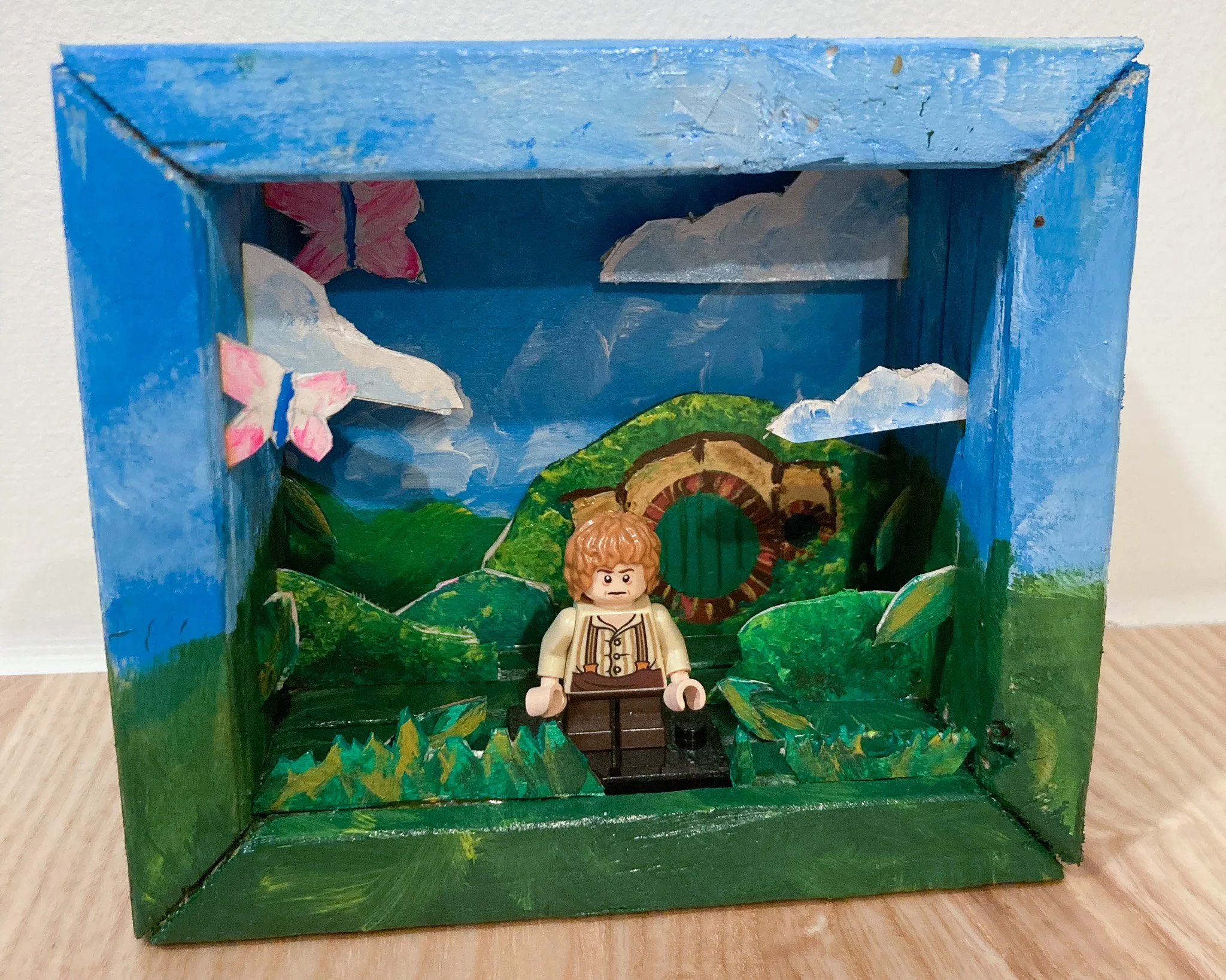 A Lego scene depicting a landscape with green hills, blue sky, pink flowers, clouds, and a hobbit hole with a round door, featuring a Lego figure with curly hair and a beige outfit standing in front of it.