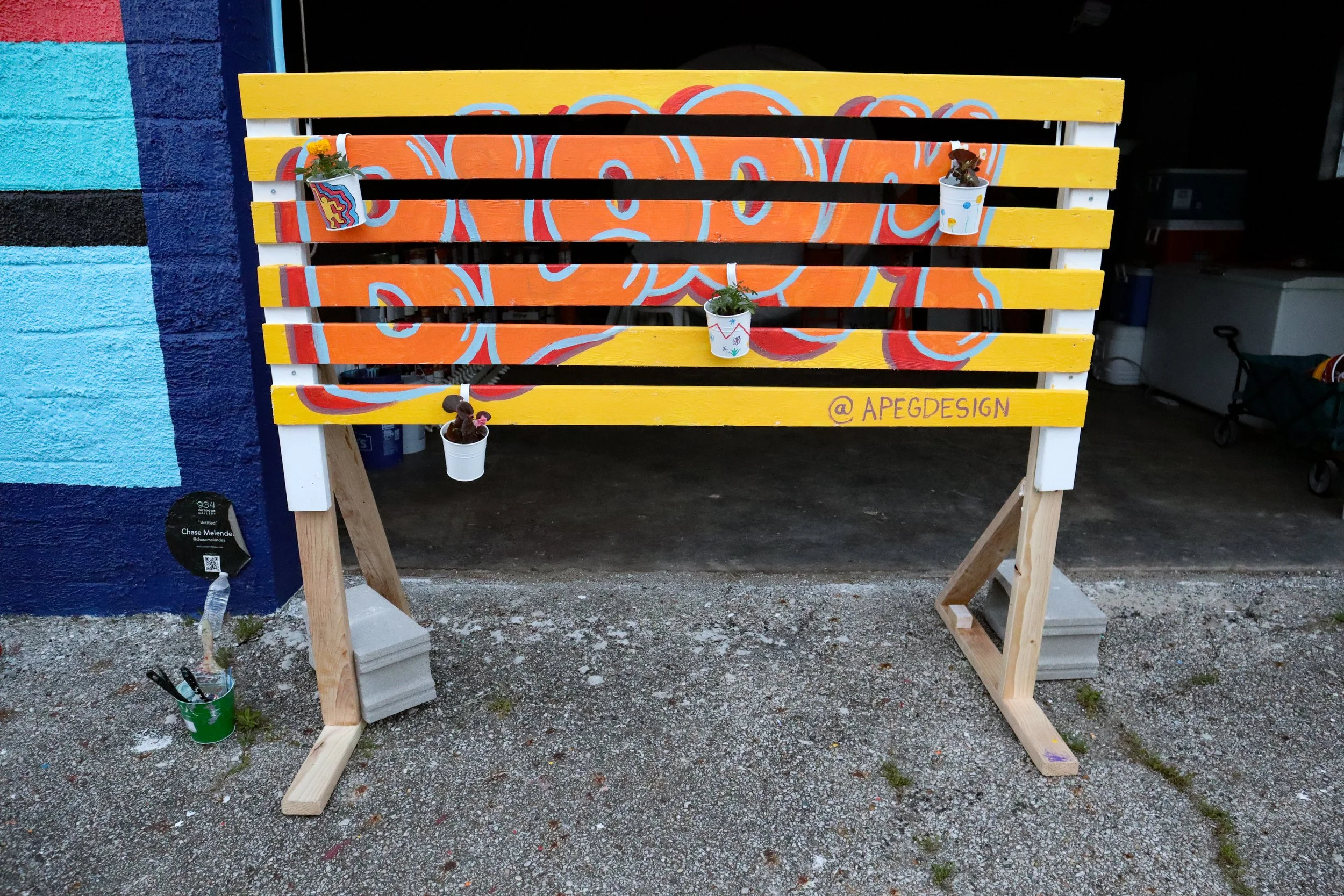 A colorful wooden fence painted with orange and yellow stripes and a pink and orange flower design in the center. The fence is supported by wooden stands and decorated with small potted plants. The background includes a blue and black wall and a garage with a shopping cart inside.