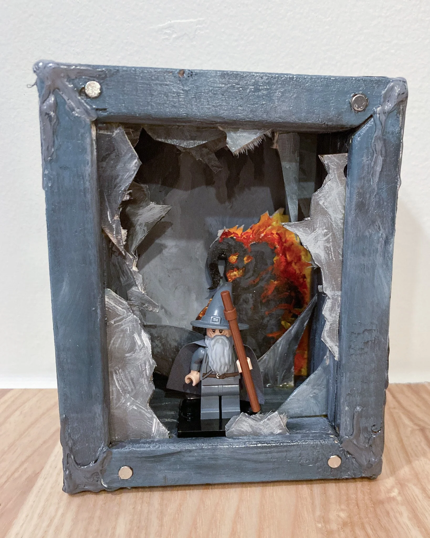 A LEGO wizard figurine with a gray hat and beard, holding a staff, standing in a decorated box with a dragon background.