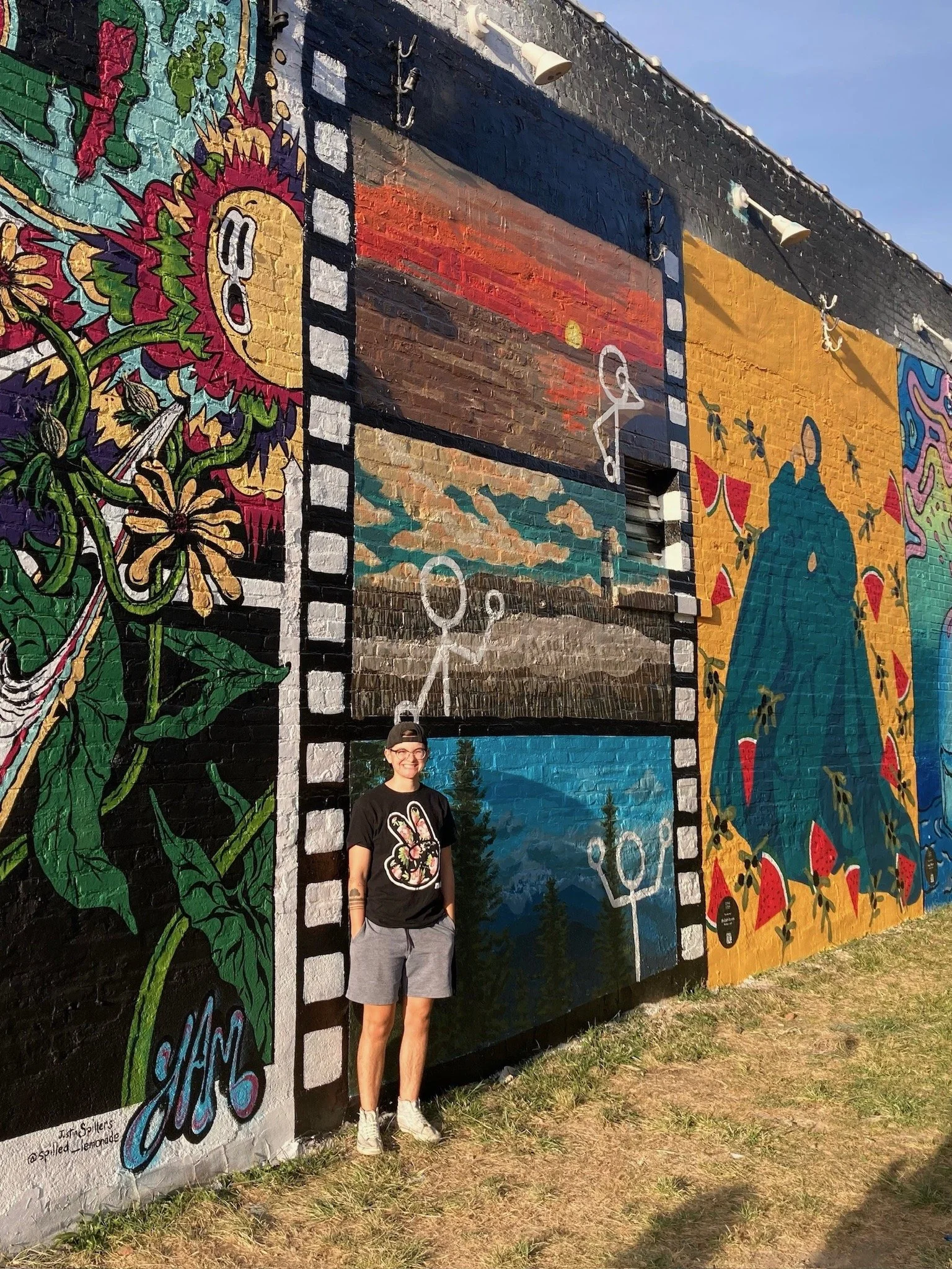 Person standing in front of a colorful mural painted on a brick wall featuring various artistic scenes and characters.
