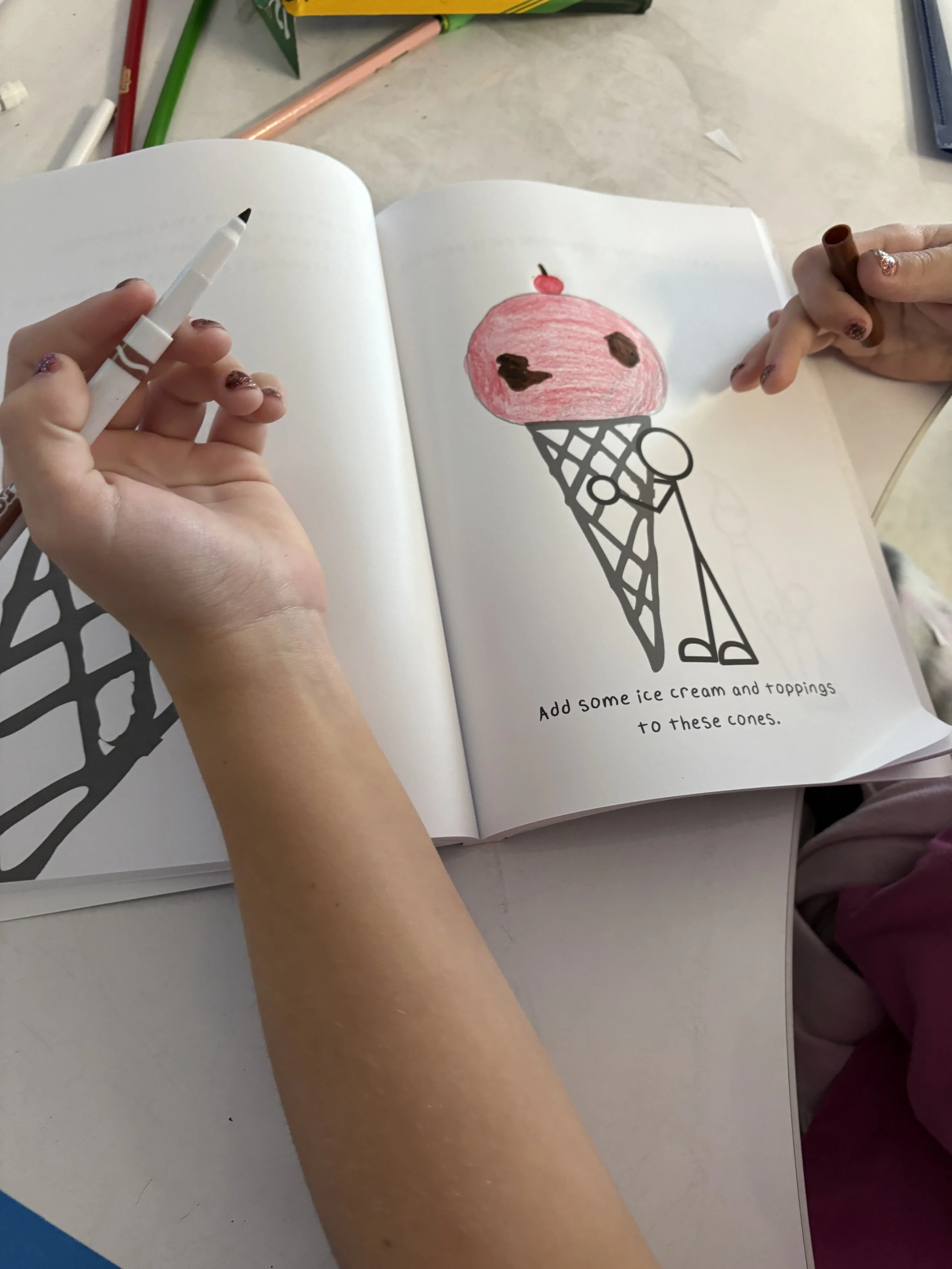A child is drawing a picture of an ice cream cone with pink ice cream, chocolate spots, and a cherry on top in a notebook. The text says, "Add some ice cream and toppings to these cones."