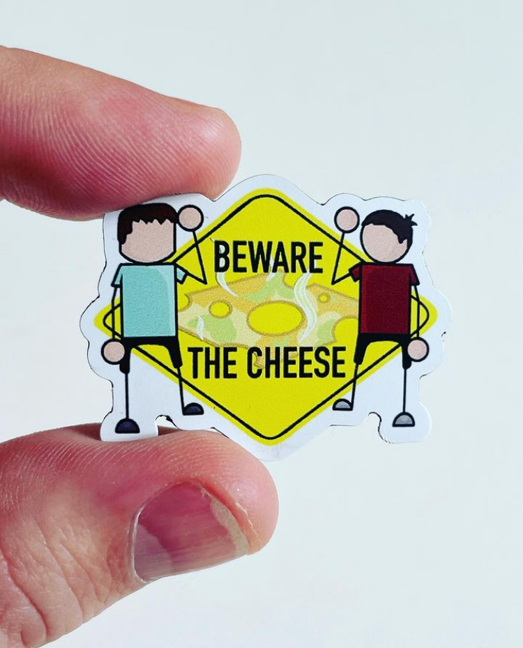 A sticker with cartoon characters holding strings connecting to a yellow cheese-shaped sign that reads 'BEWARE THE CHEESE'.