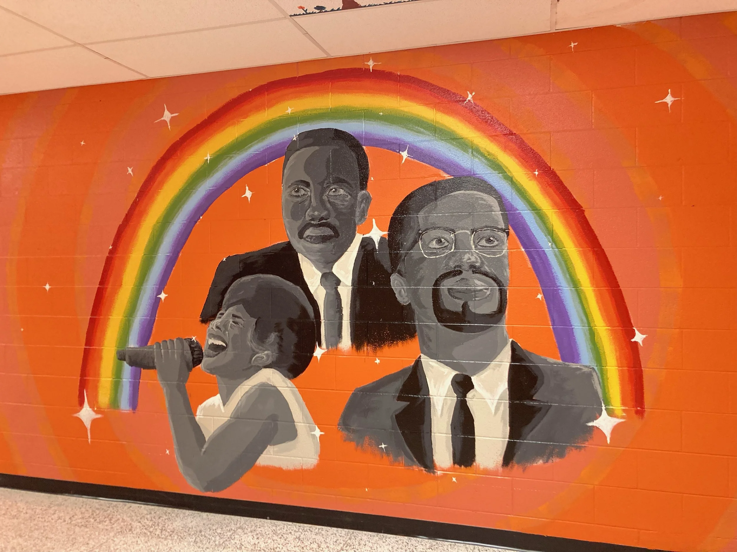 Wall mural depicting Malcom X, Martin Luther King Jr, and Aretha Franklin in grayscale with a rainbow and stars in the background.